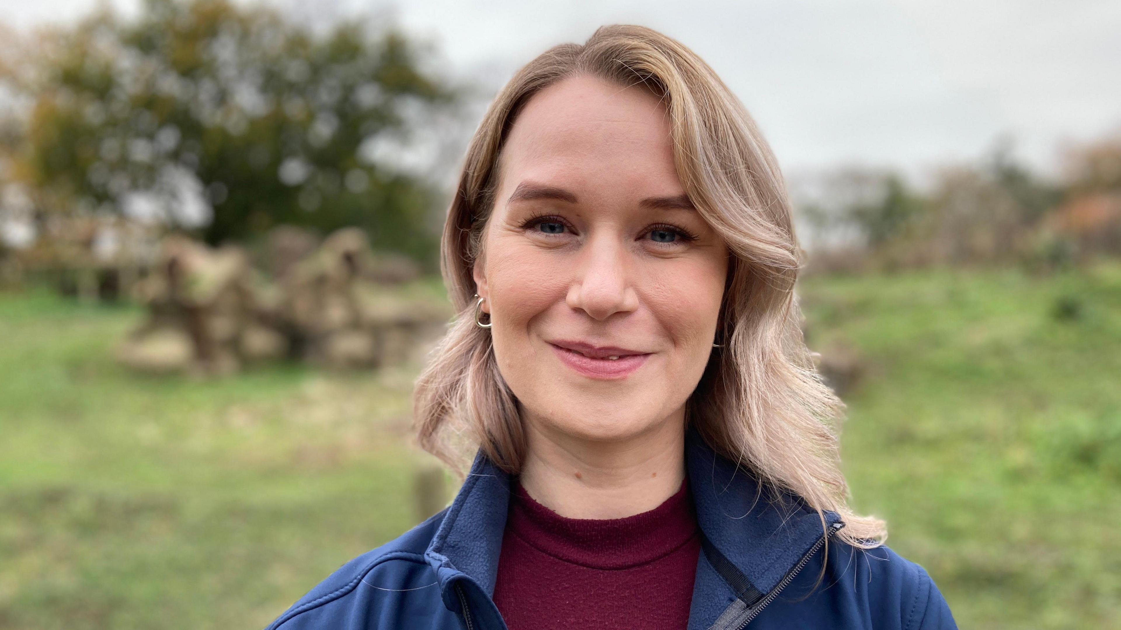 Headshot of Sarah Richdon. She has pale skin and blonde hair, and is wearing hooped earrings and a burgundy top. She is smiling slightly at the camera.