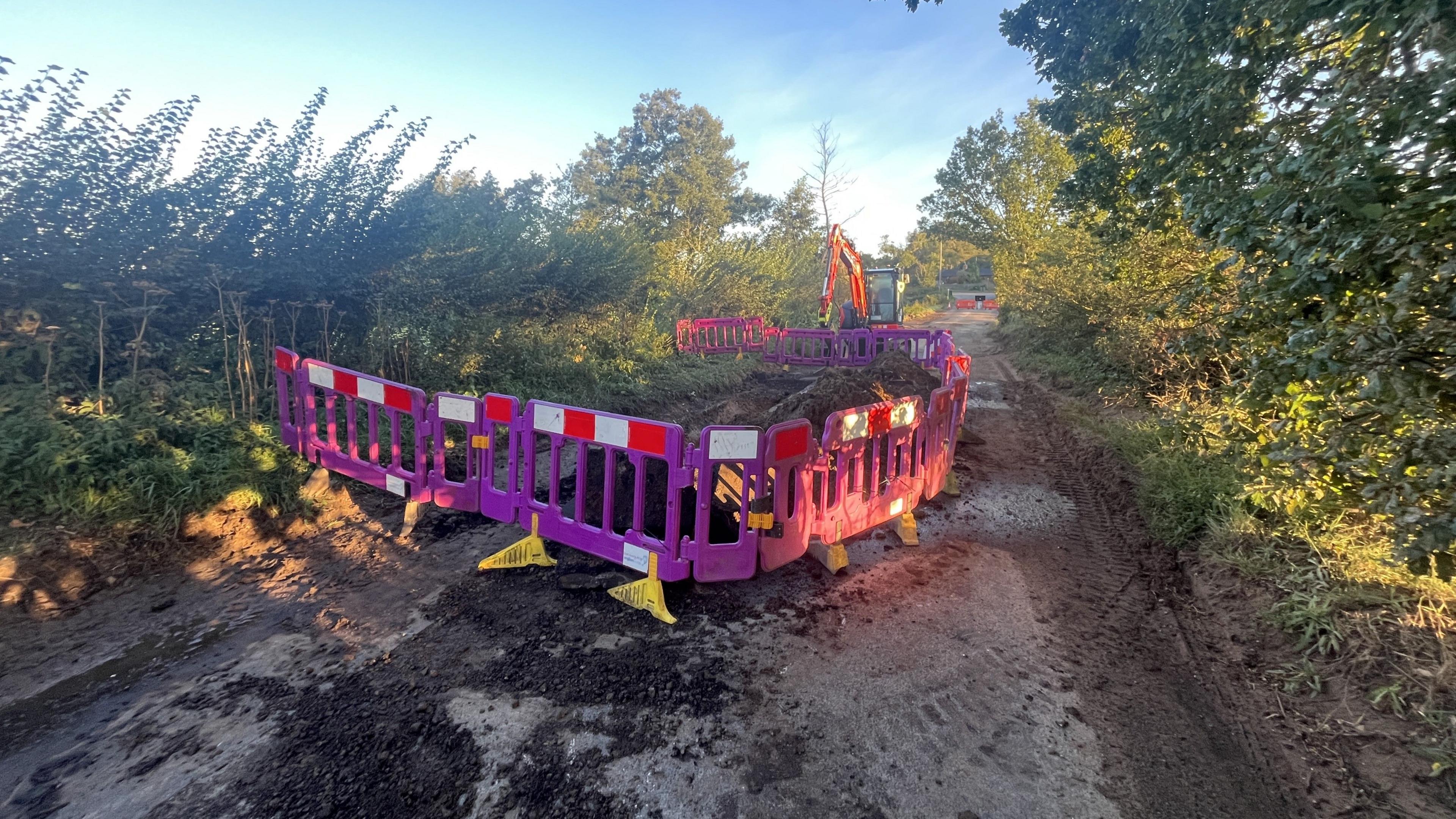 A section of a single track road is blocked by purple barriers and a digger can be seen parked behind it.