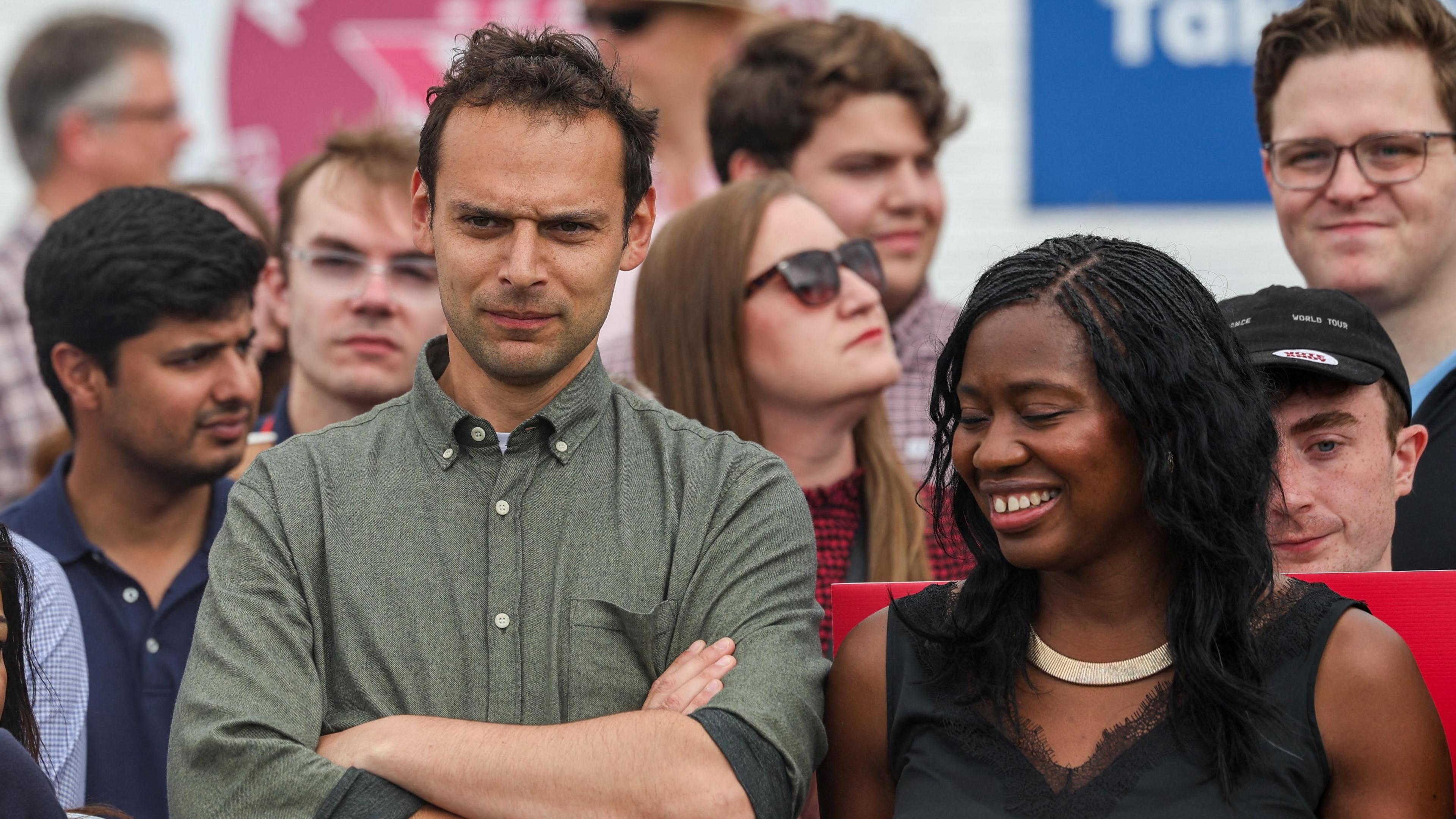Josh Simons, in a green shirt with a button down collar, folds his arms and looks on as Burnham delivers a speech to supporters. A smiling Miatta Fahnbulleh, in black top, is stood next to him 