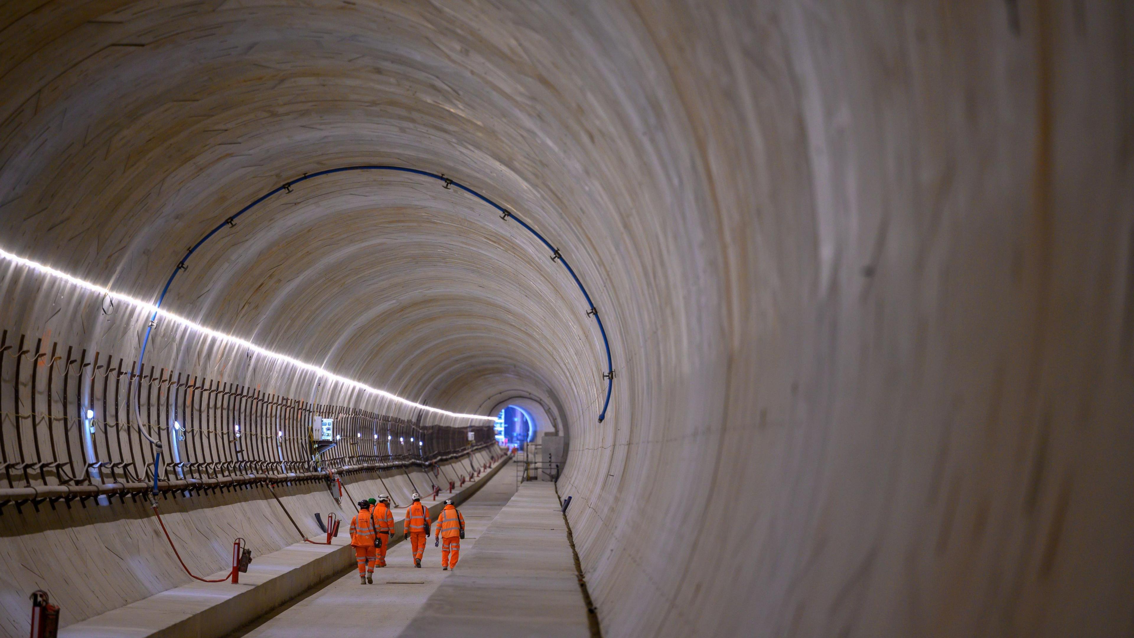 A large long tunnel with a group of four people dressed in fluorescent orange and hard hats walking through