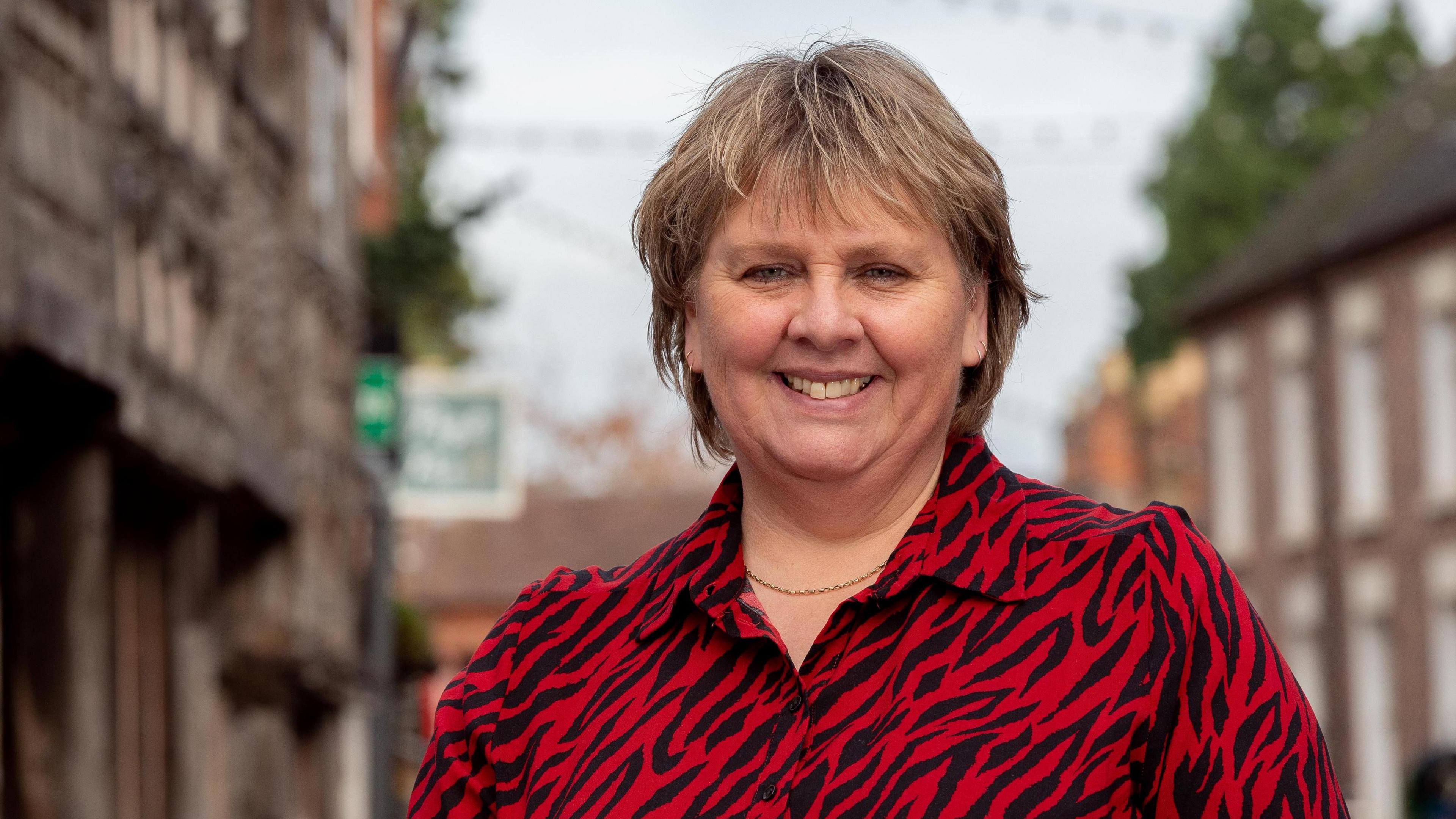 Woman with short light brown hair is looking at the camera and smiling. She is wearing a red and black animal print blouse and a gold necklace. There is a blurred high street behind her.