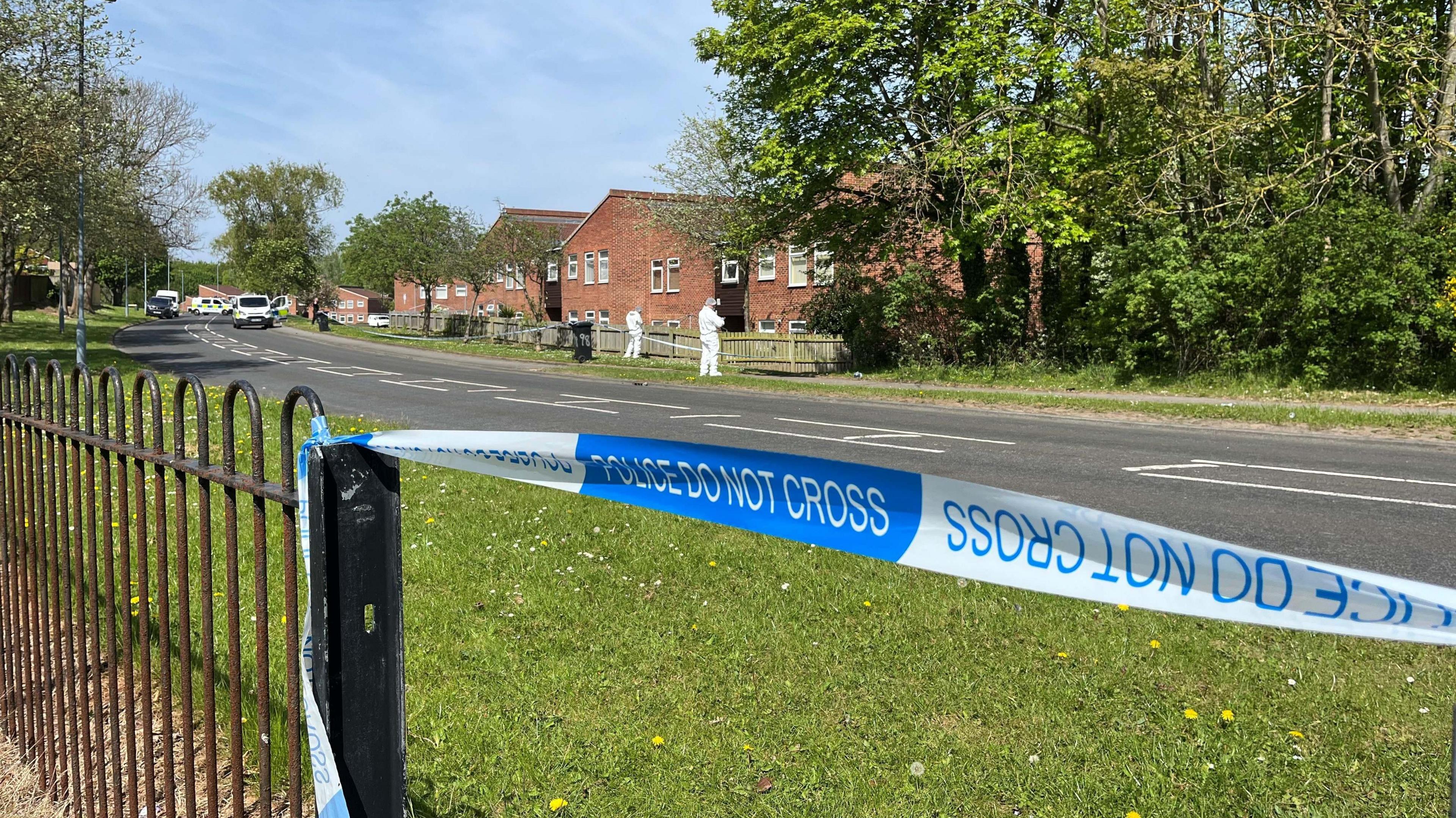 In the foreground, blue and white police tape has been attached to a black fence. In the background, forensics officers in white suits are inspecting ground near a fence with a row of houses behind.