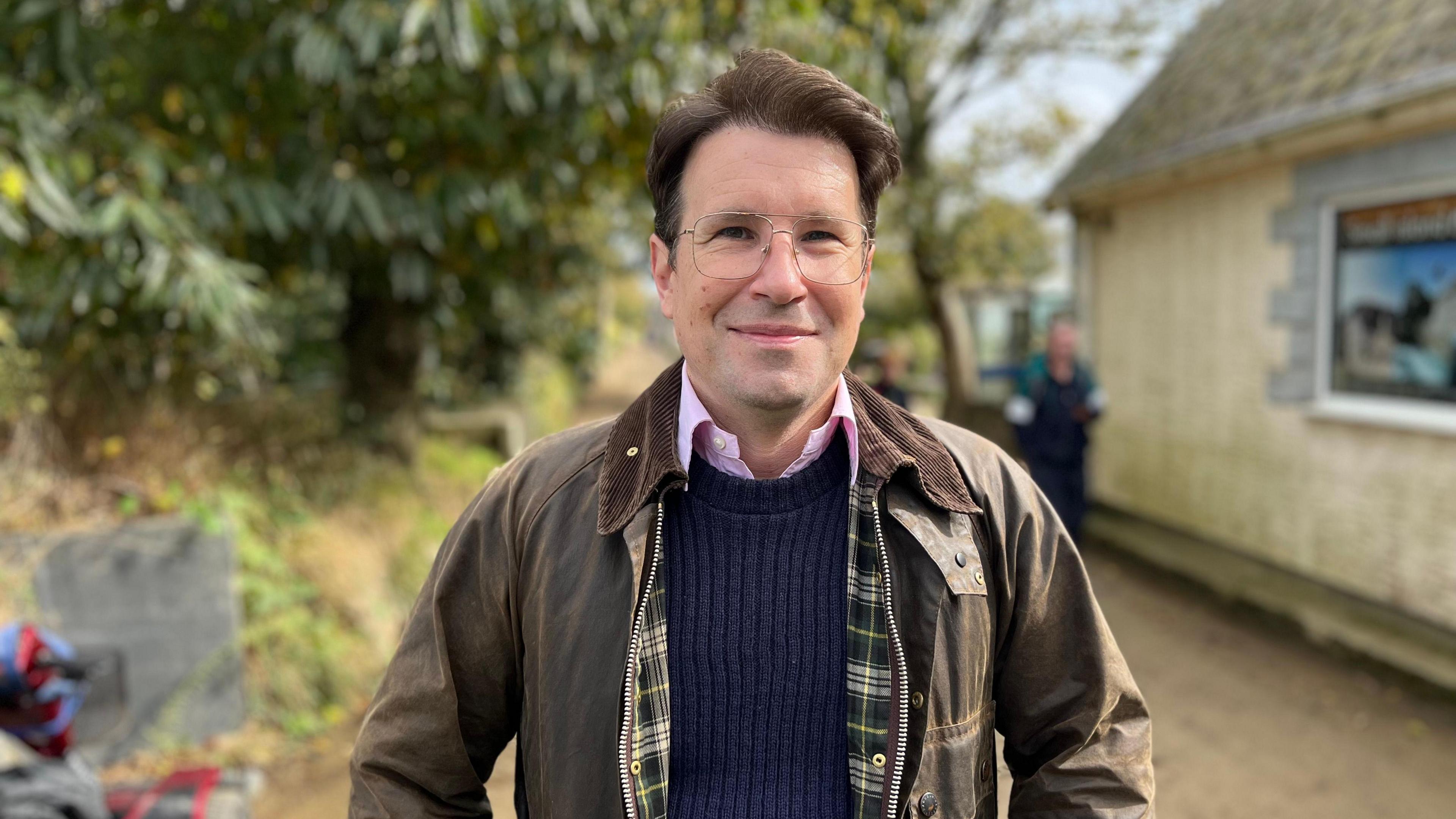 A man with glasses smiling towards camera. He is wearing a green jacket and a blue jumper. 