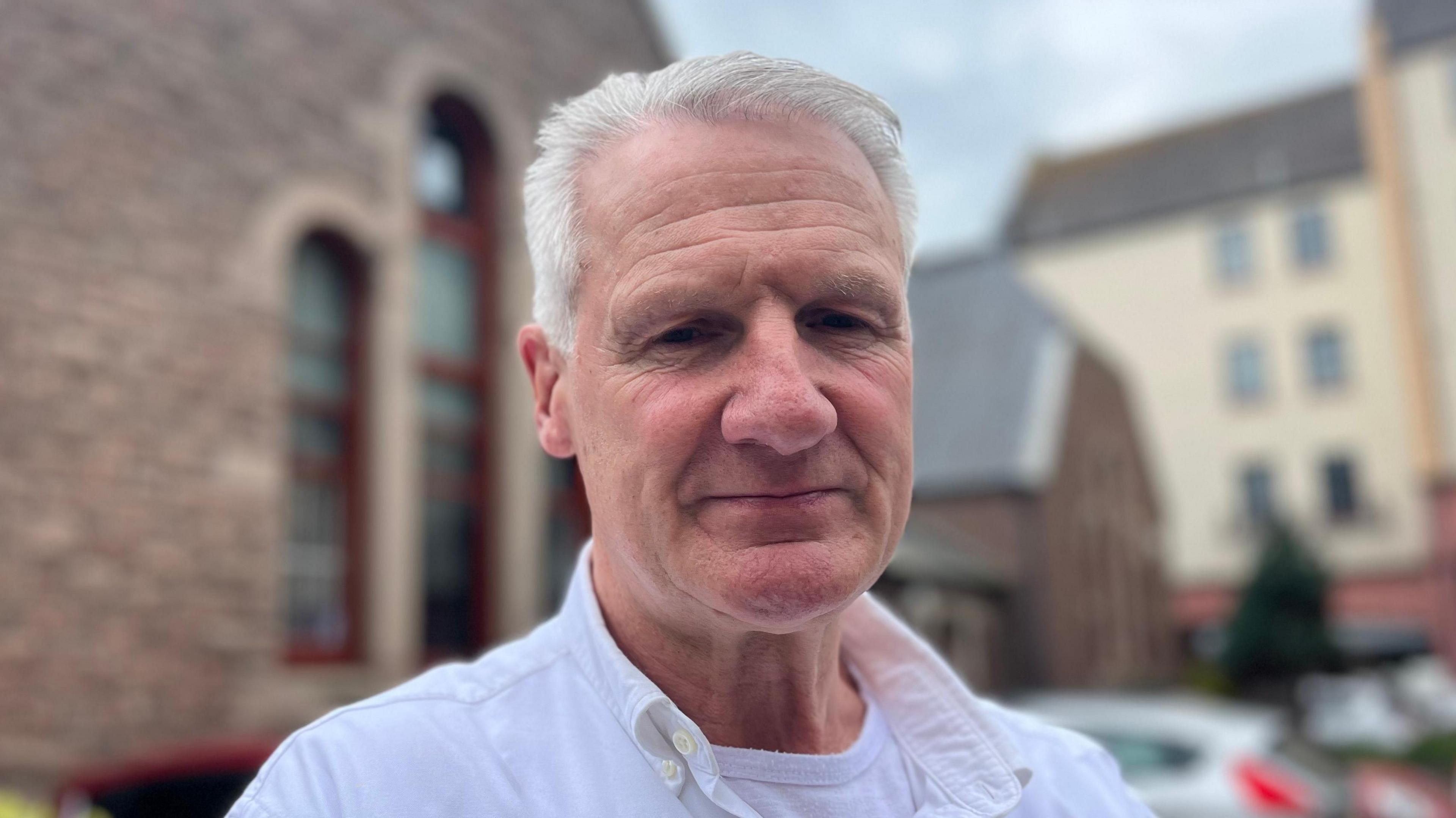 Deputy Tom Binet is a white man with white hair wearing a white collared shirt over a white vest. He is staring down the camera lens. The background is an out of focus church hall behind him.