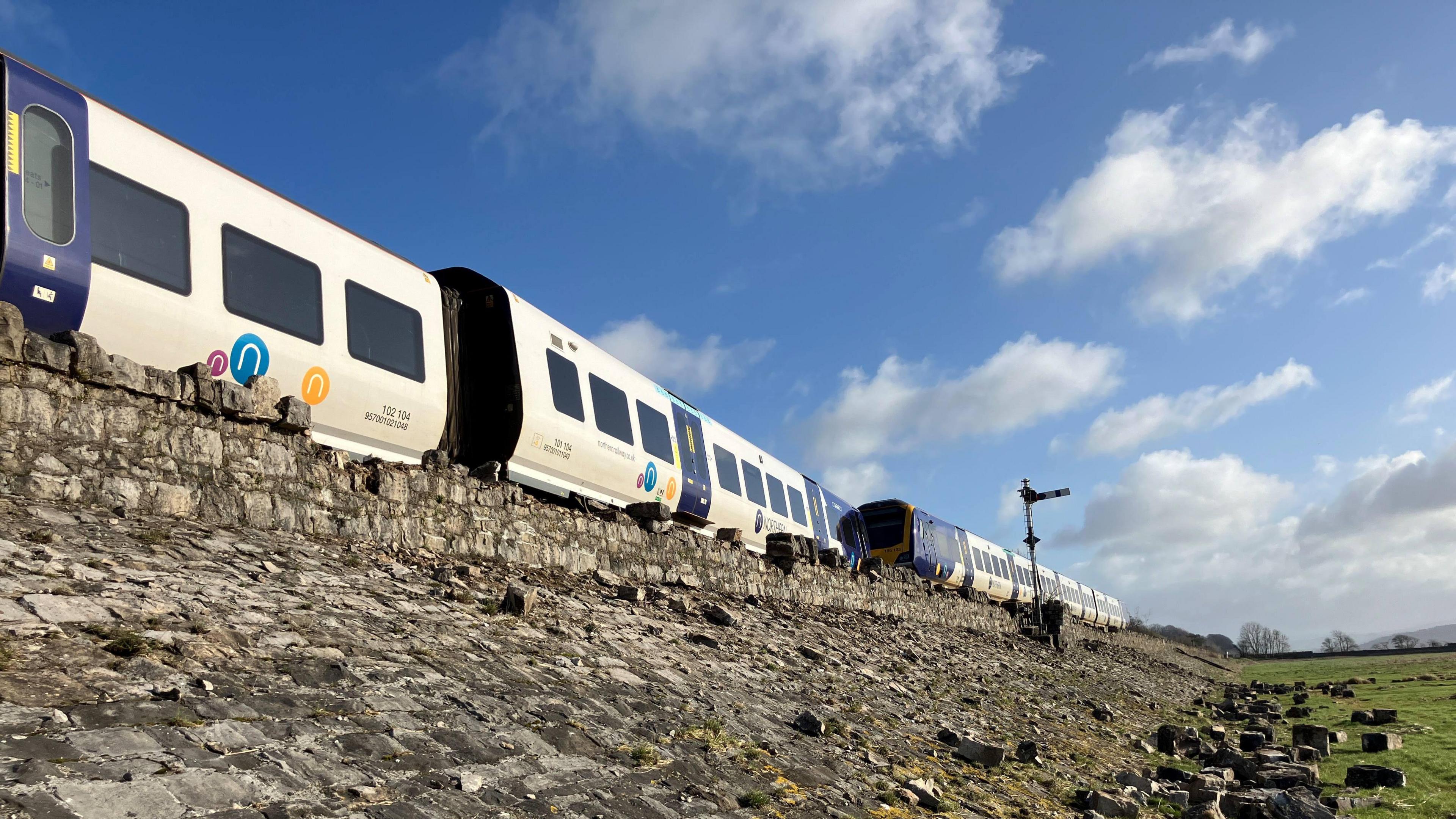 Three carriages of the derailed train are visible on the tracks above a rocky embankment. The derailed section of the train is not visible