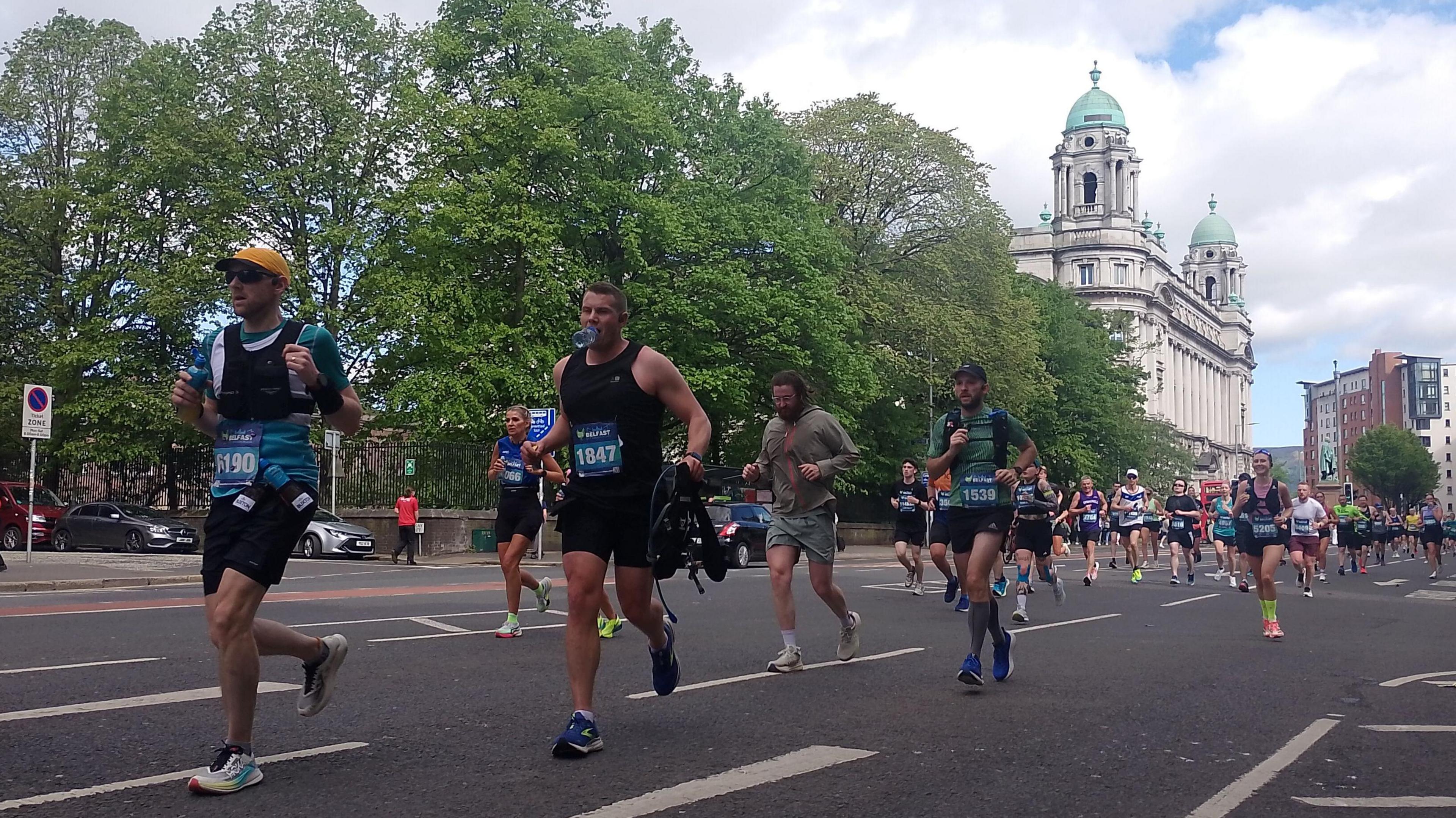 Numerous runners are captured wearing different coloured sportswear as they run along a street. In the background is green trees and a stone building.