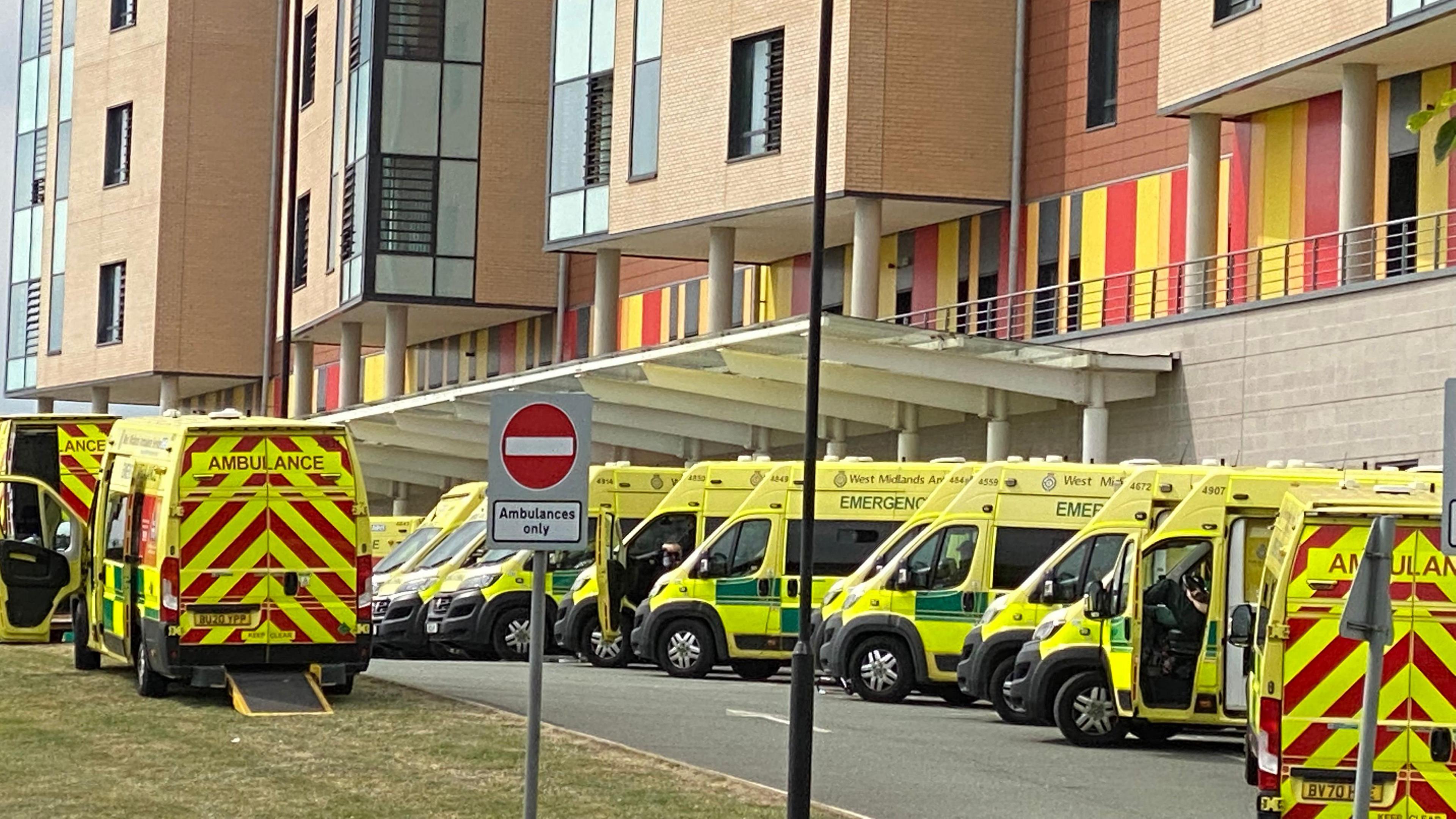 A number of ambulances parked up in a line outside the Royal Stoke University Hospital, most on a road, with one on grass, where a no entry sign is in the foreground