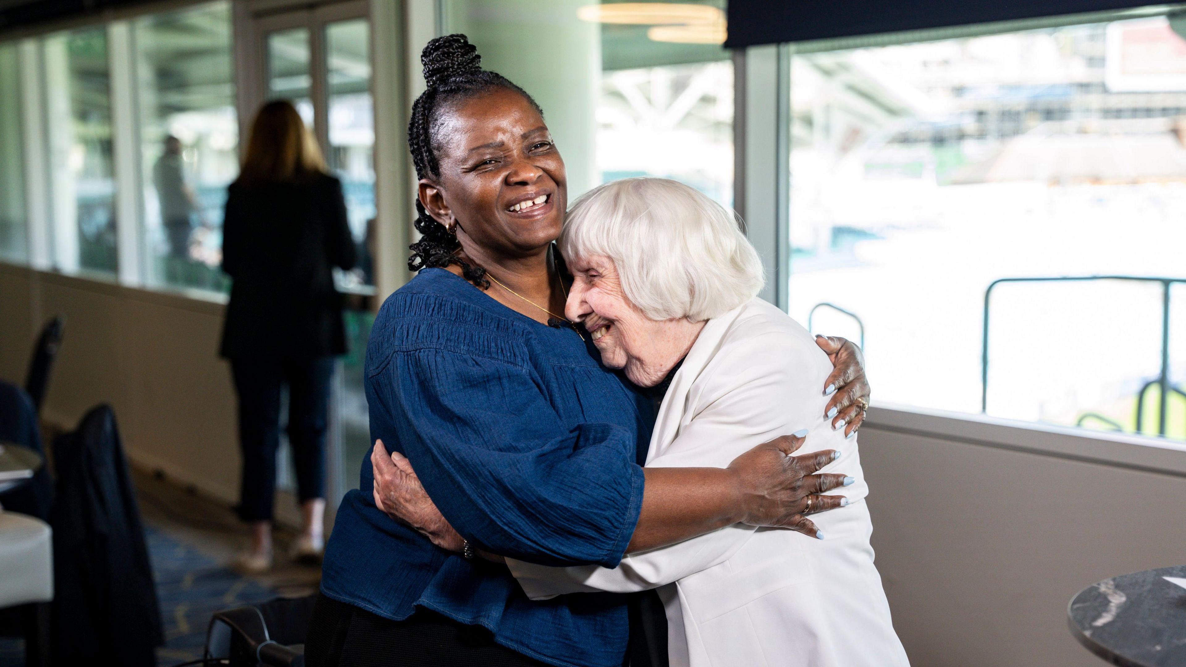Teju Adedayo hugs Betty Brown. Ms Adedayo has dark hair styled in braids and is wearing a blue top. Ms Brown has grey hair and is wearing a white jacket.