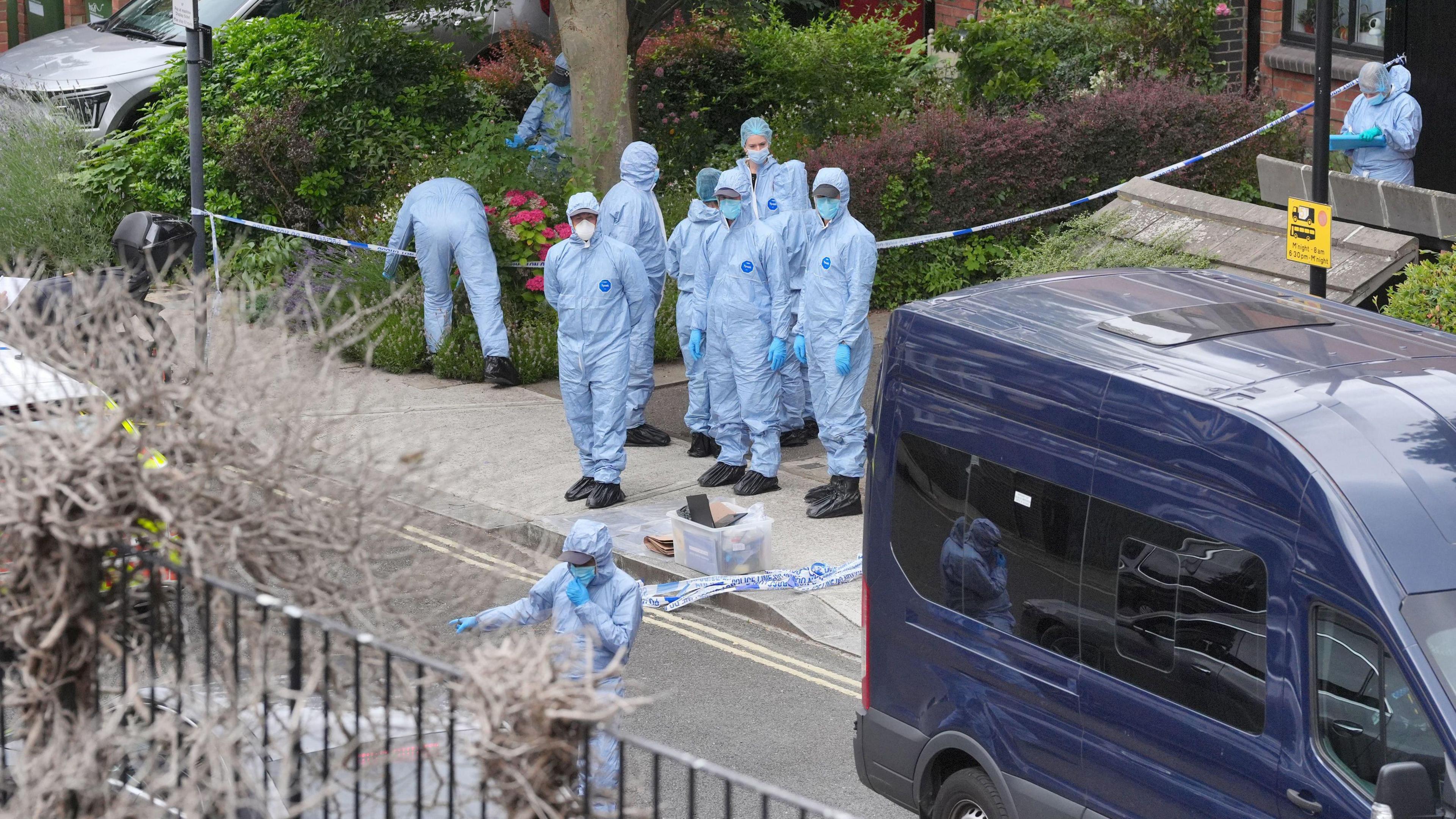 A group of people wearing forensics suits. The suits are blue and the people are wearing surgical face masks and latex gloves. Police tape can be seen too.