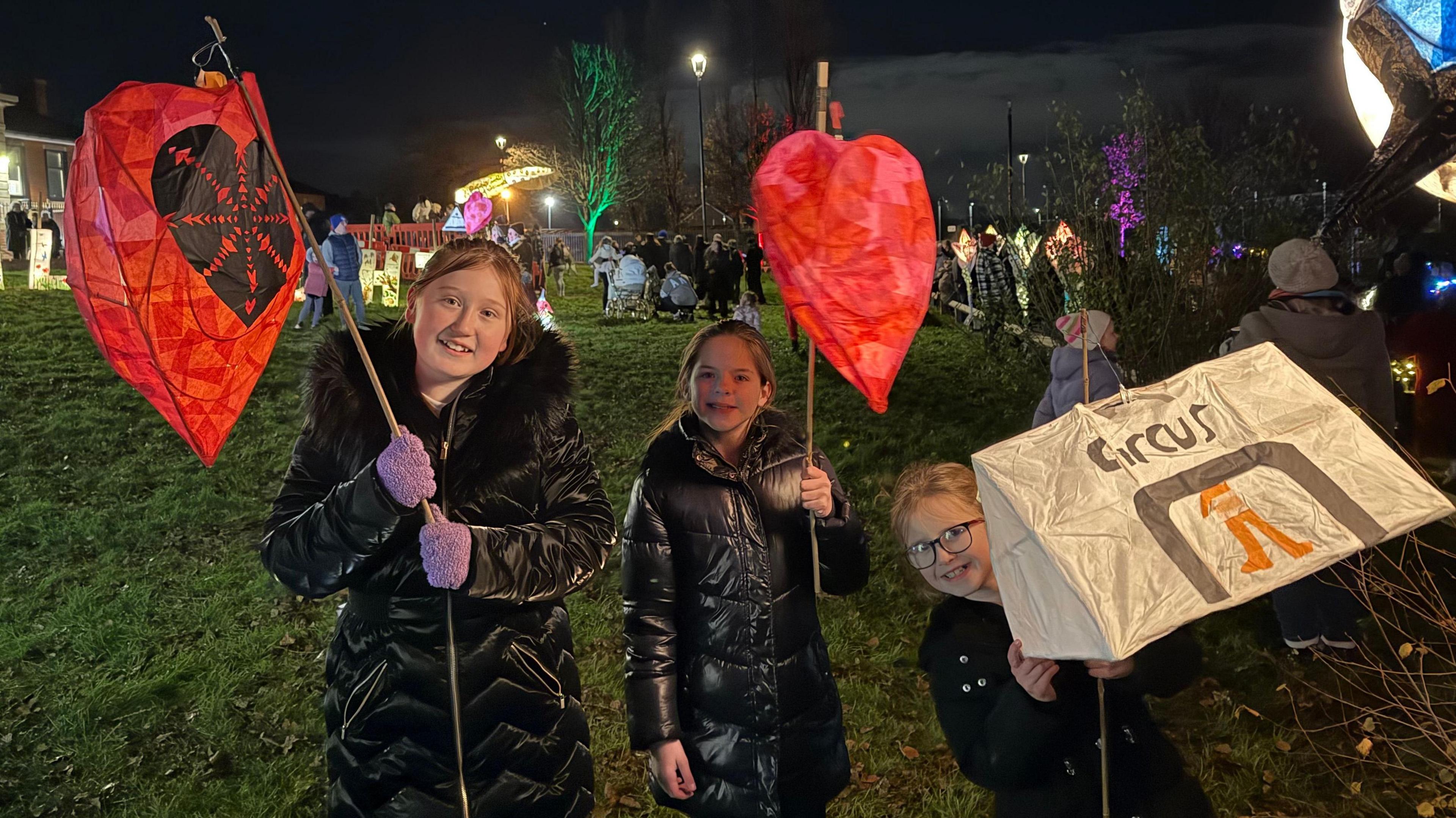 Three young girls hold lanterns, two shaped as red hearts and one as a circus tent.