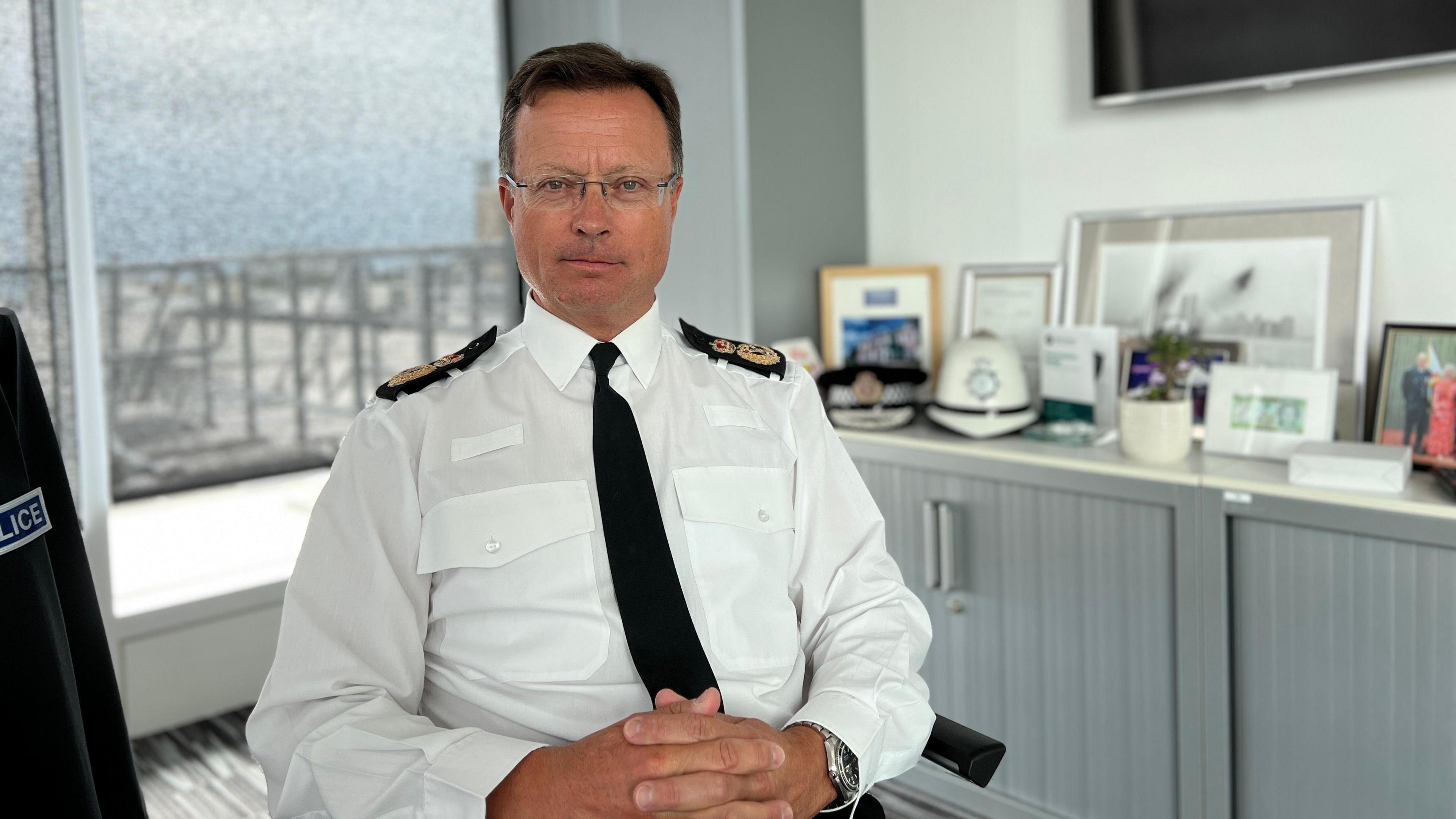 Chief officer Robin Smith - a man with a straight line smile, rectangle non-rimmed glasses, silver watch on his left wrist, dark brown hair wearing a white long sleeve shirt with a black tie and epaulets, hands crossed in his lap, the background is blurred of an office with a large window behind Mr Smith 