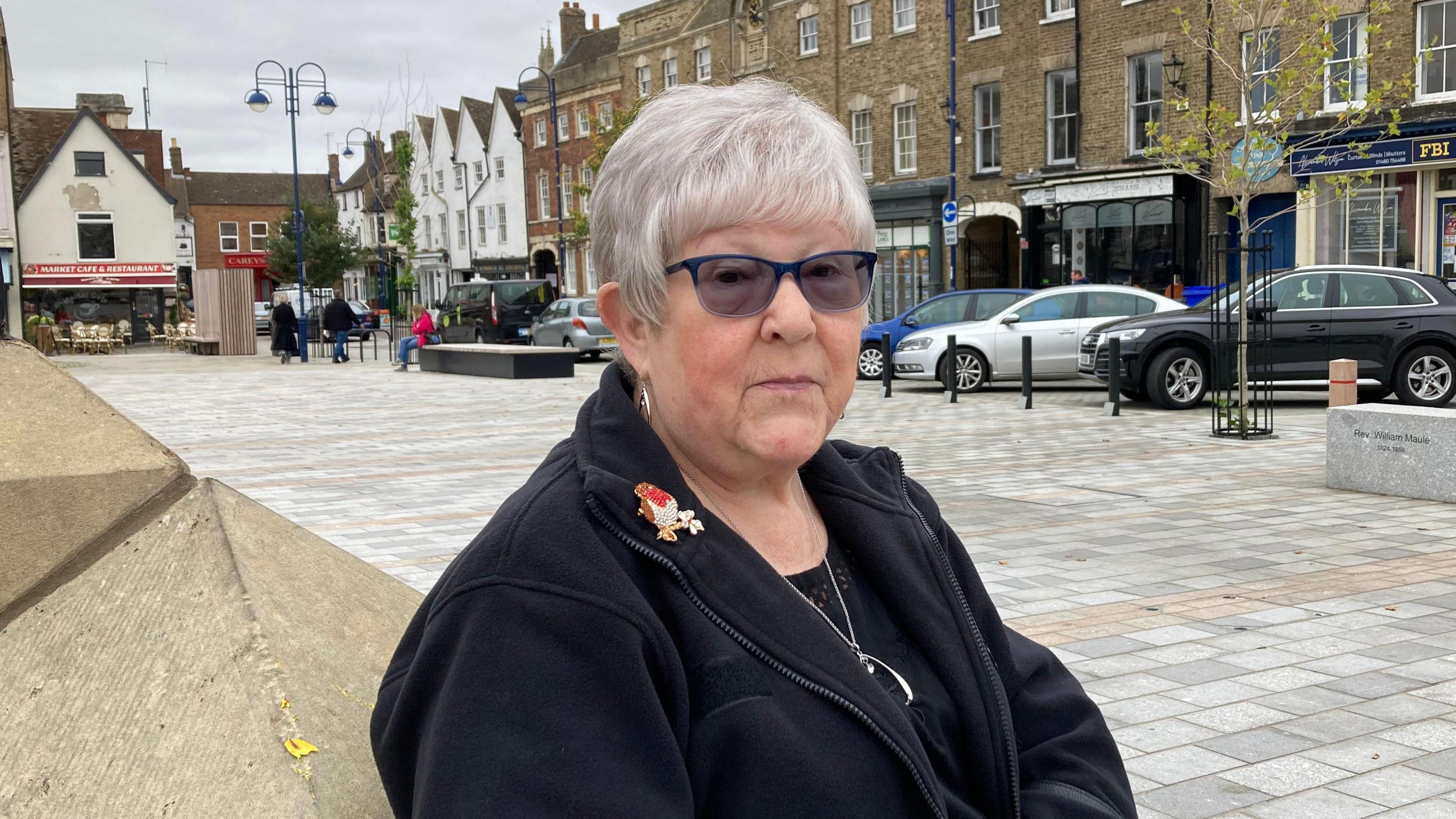 Anne Hill sits on a seat in a pedestrian square. She wears a black jacket, dark glasses and a sparkly robin brooch. In the background are some parked cars and some shops.