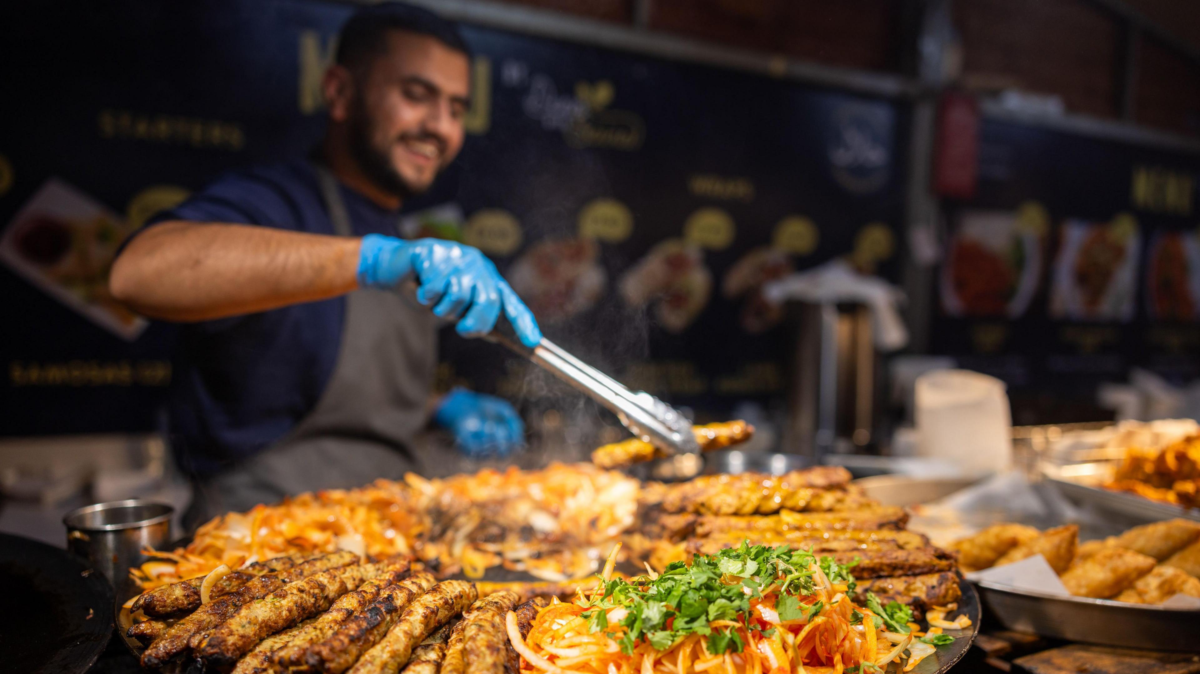 A market trader wears an apron and blue rubber gloves to cook meat skewers.