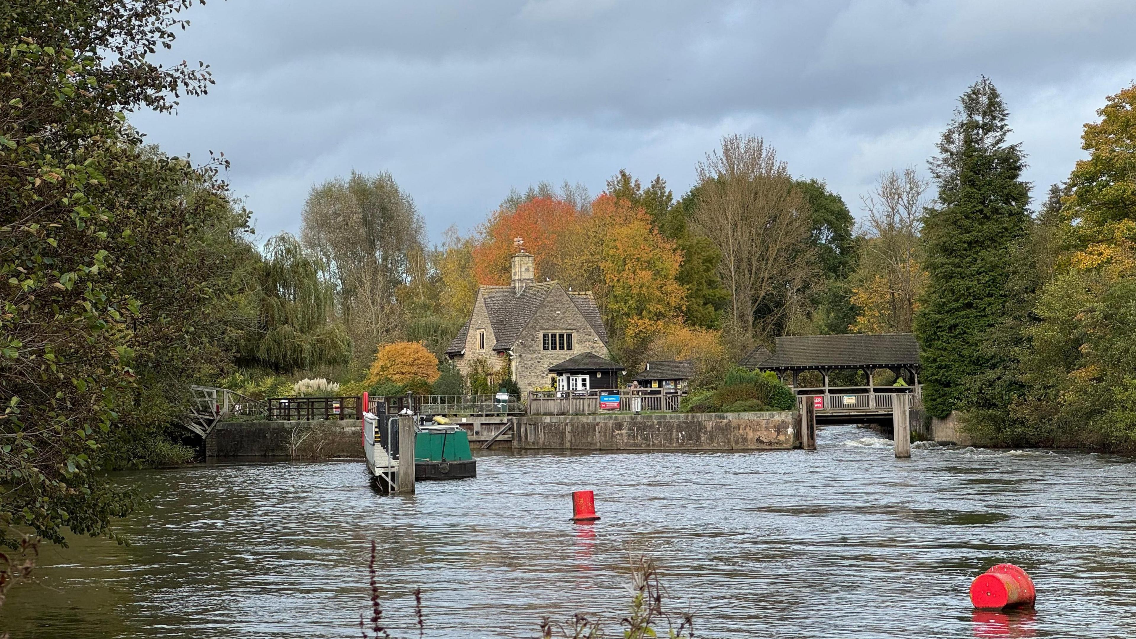 A stone building sits on an island between two bodies of water. There is a green canal boat in the middle of the water. The banks of the river are lined with trees, some of which are starting to change colour. There are two red buoys in the water.