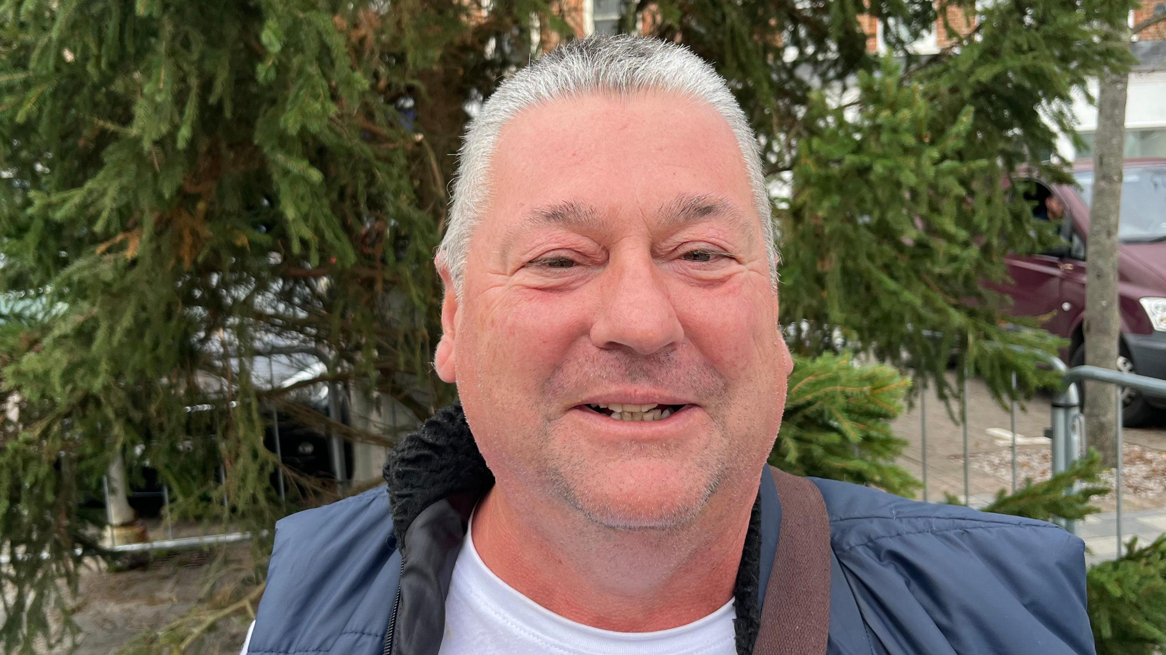 A man smiling at the camera, standing in front of the lower branches of a Christmas tree. He is wearing a blue puffy gilet and grey tshirt, and has grey hair.