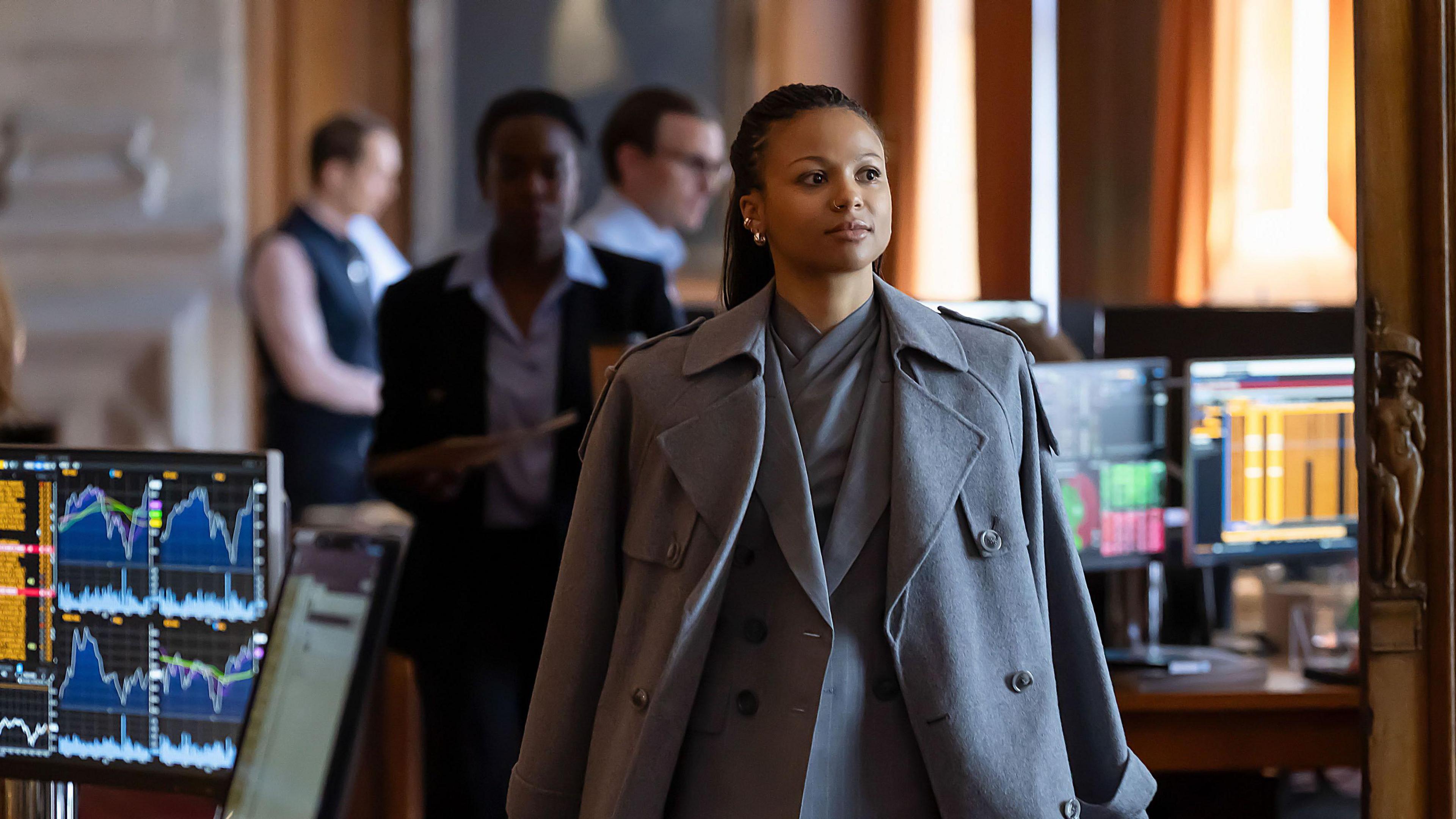 Harper, played by US actress Myha'la, wearing a grey coat in an office scene with computers behind her