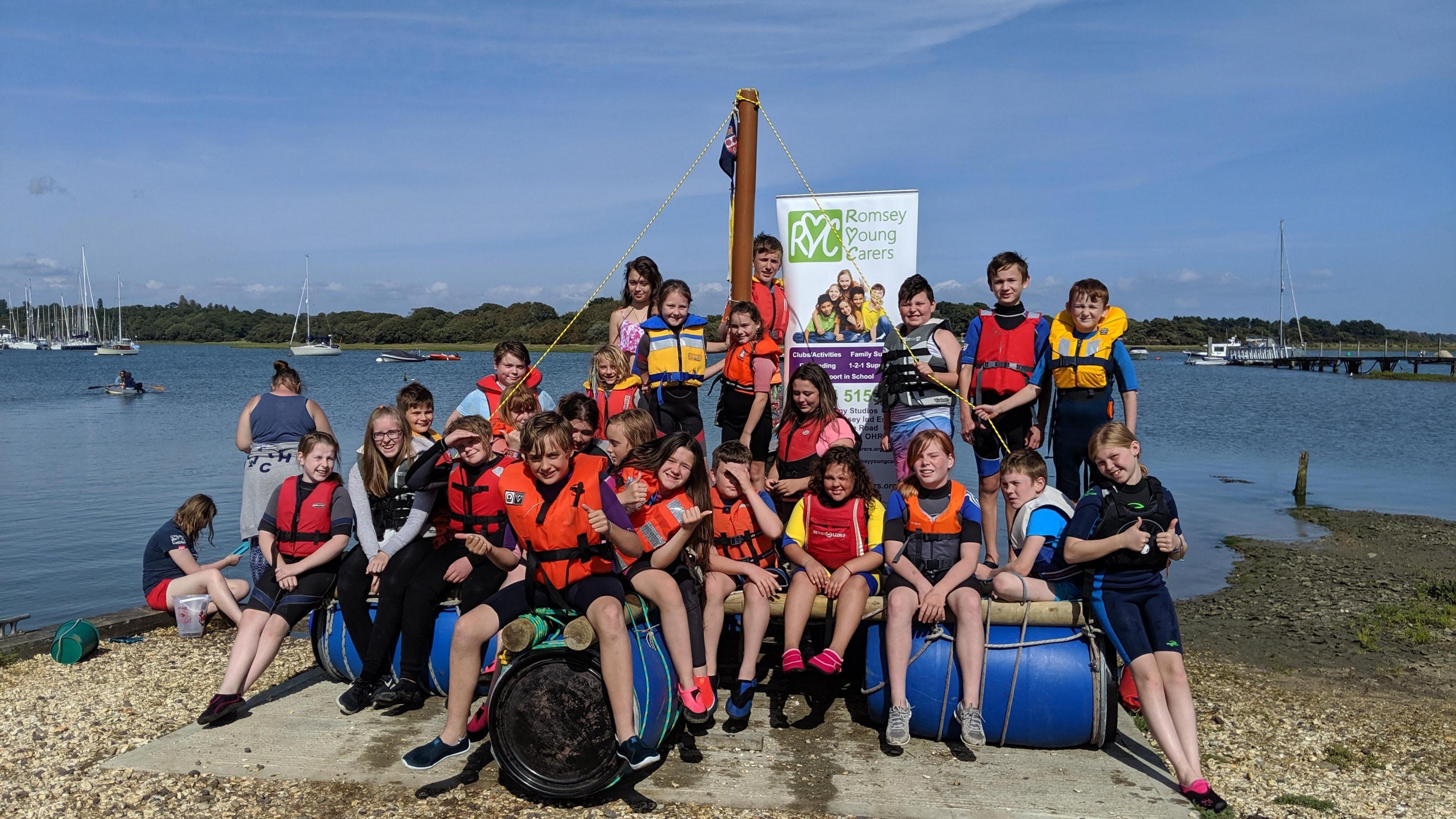 A group of children sat on makeshift boat equipment next to a lake during a Romsey Young Carers activity day