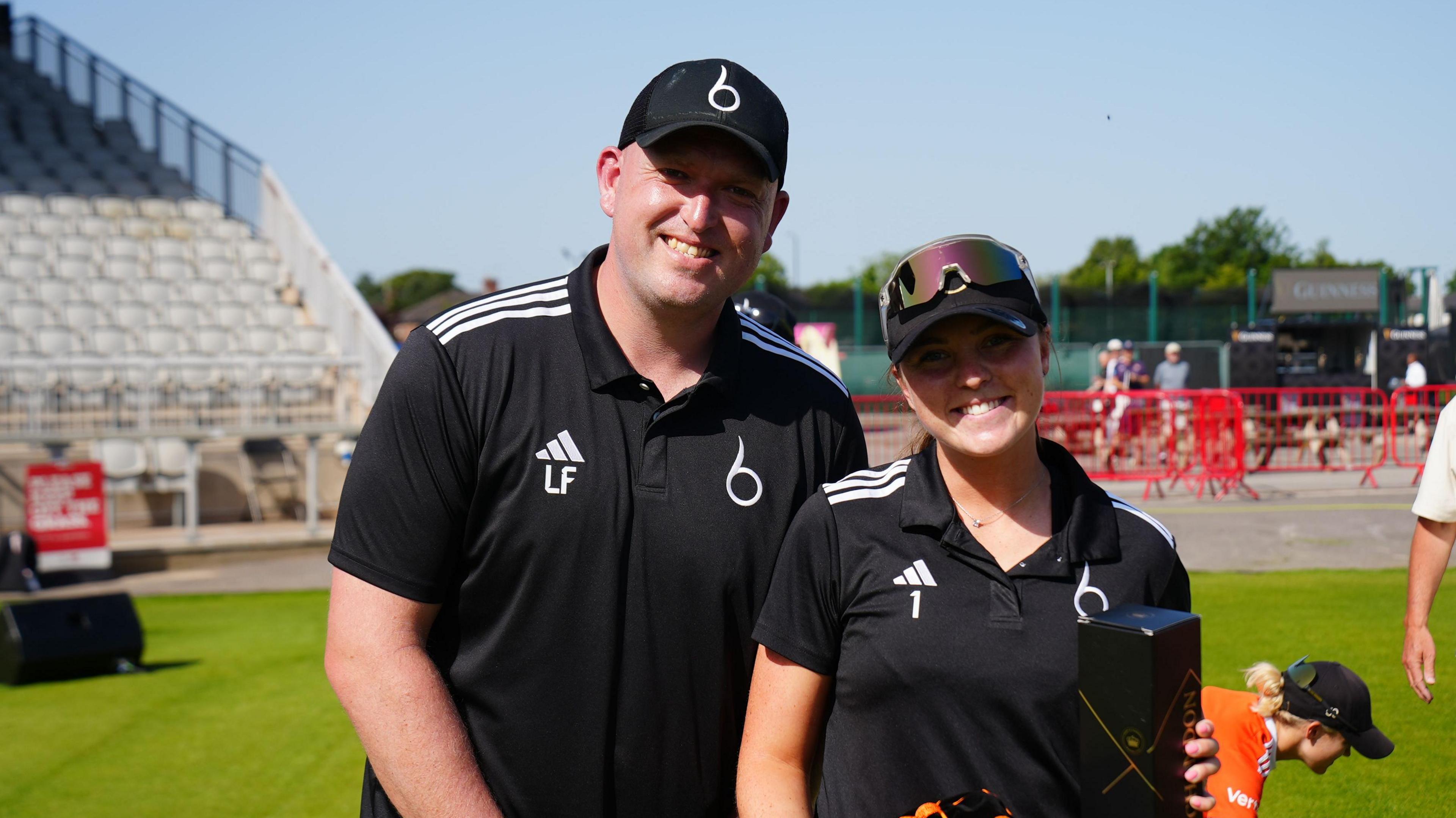 Luke Fletcher and Grace Ballinger smile to celebrate Grace Ballinger's 100th cap at Emirates Old Trafford. It is a sunny day and they both wear black Blaze polos