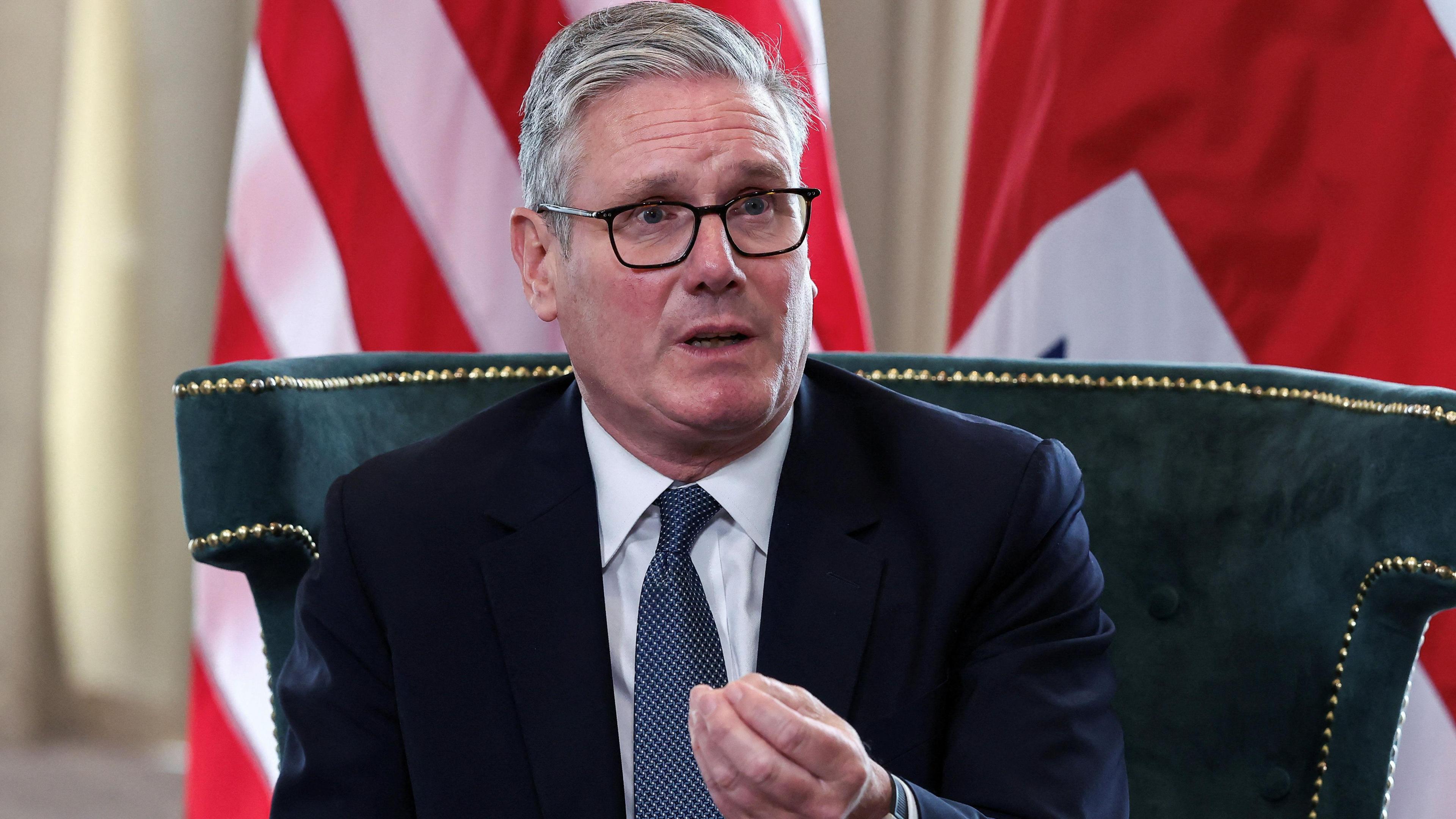 Sir Keir Starmer, sits on a green upholstered chair with gold trim, wearing a dark suit, white shirt, and blue patterned tie. He gestures with his right hand.