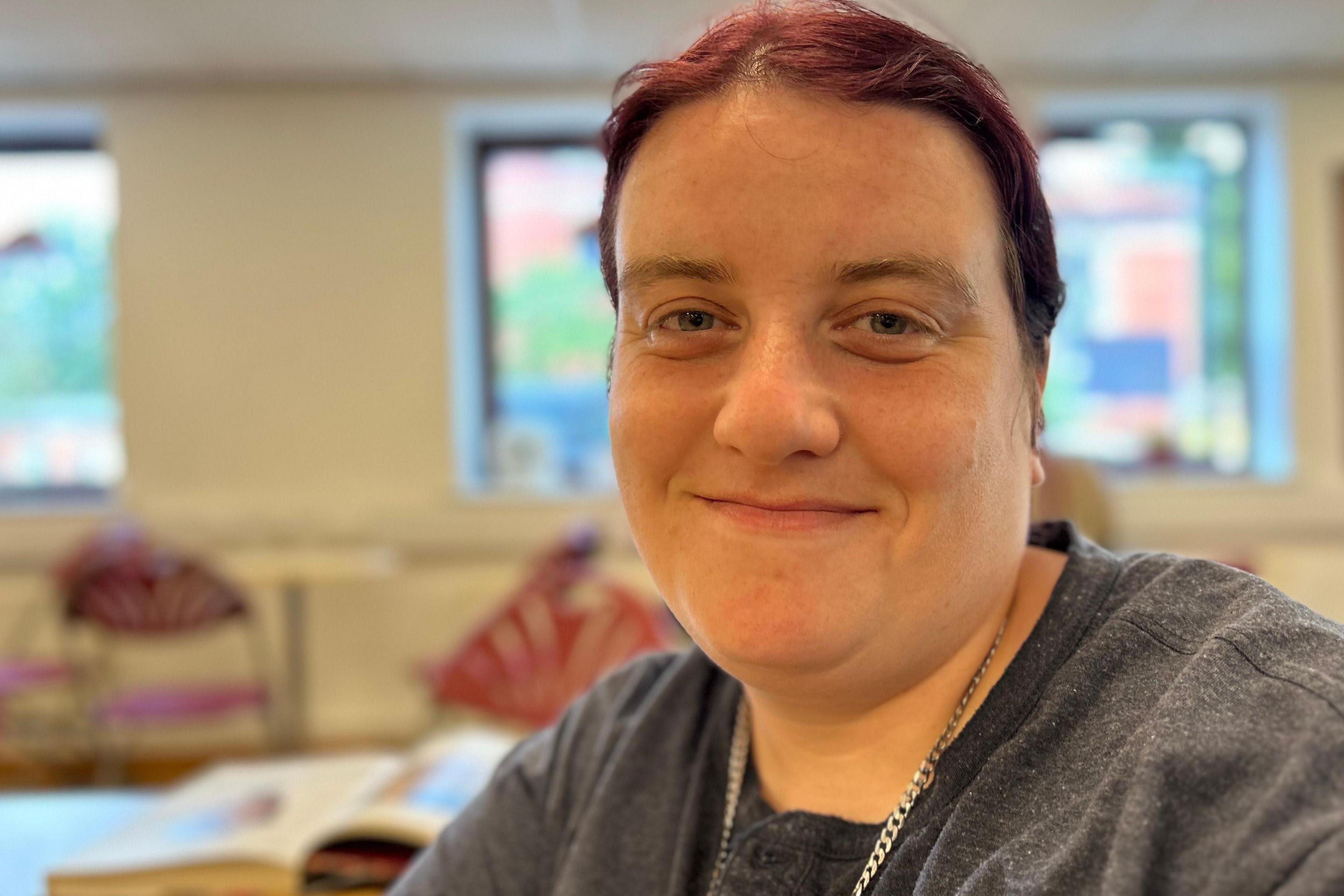 A close-up of a smiling woman who has tied-back maroon-coloured hair. She is wearing a grey T-shirt and silver necklace and sitting in a community space within a modern church hall.