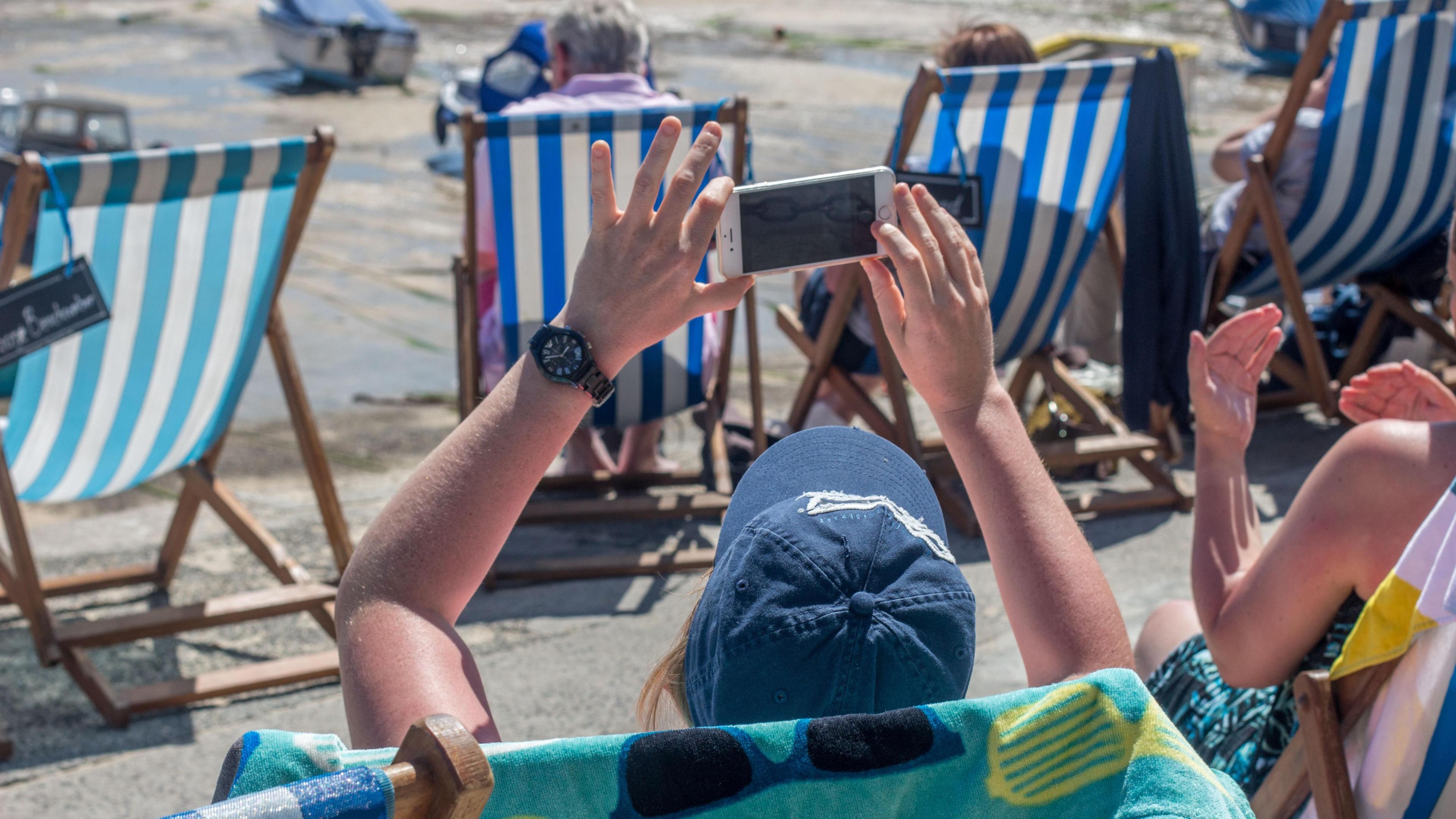 A woman sits in a striped deck chair on a beach in Cornwall. She is holding up her mobile phone to take a picture. There are more deck chairs in the background, along with small boats sitting on the sand. 