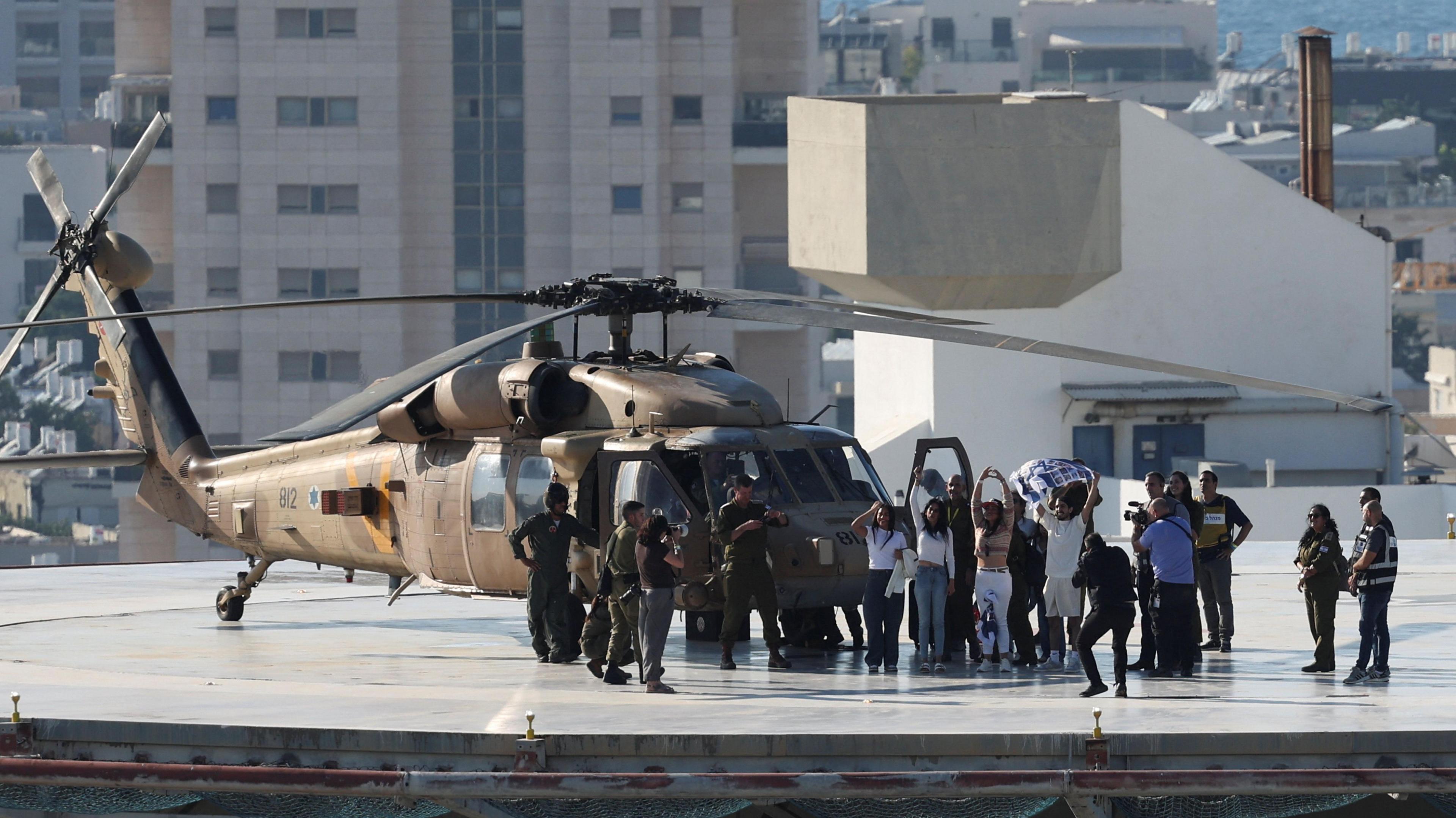 A military helicopter is on a helipad on a roof of a building. Next to it are military officials and a group of people waving and holding their arms in their air. One of them is holding a flag