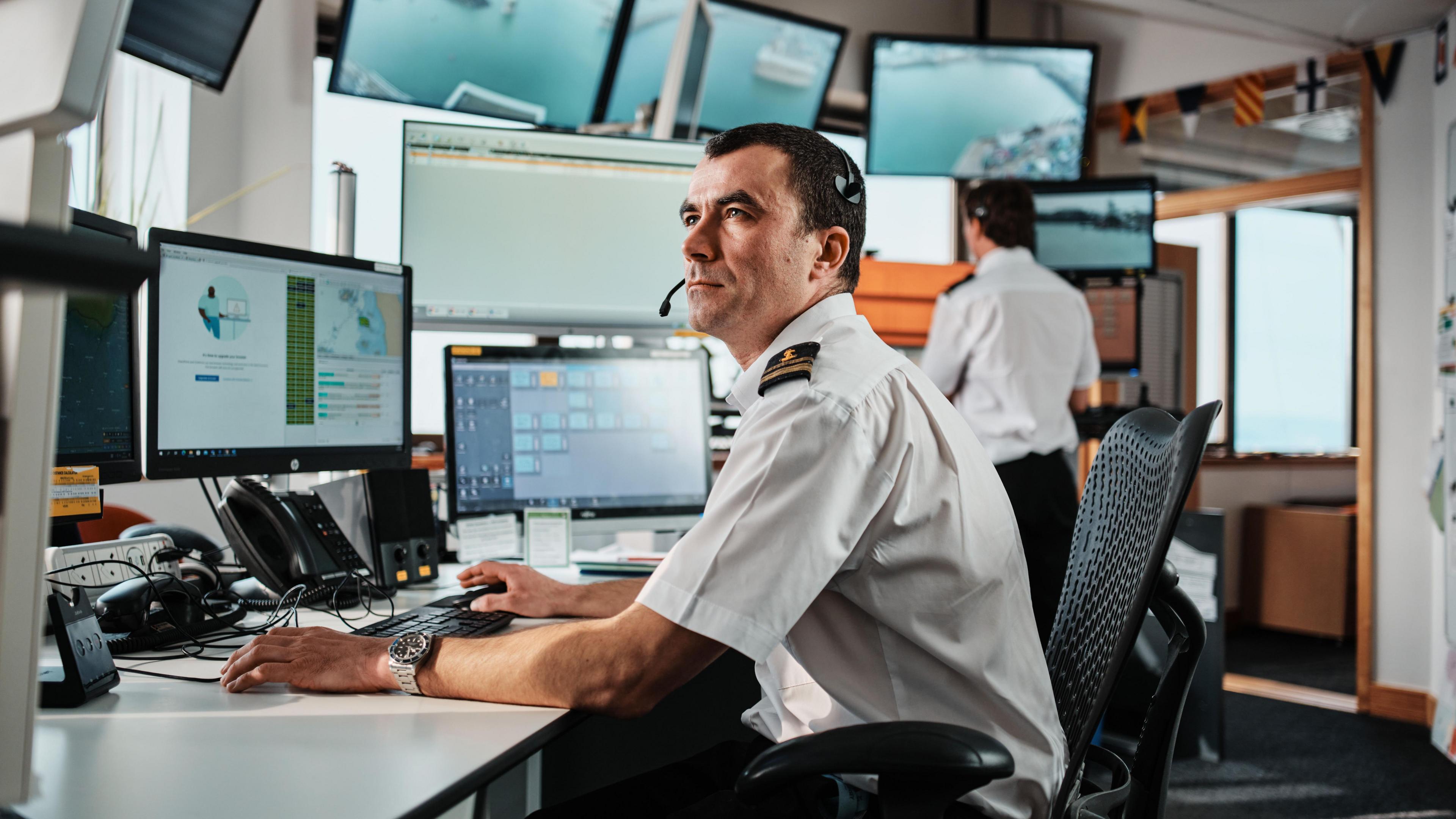 A Senior Watch Officer on duty as coastguard, with a colleague in the background working as a Vessel Traffic Services Officer. There are computer screens on a desk, along with telephones. The setting is the Maritime Operations Centre in Maritime House, which is the location for the Open Day on Sunday.
