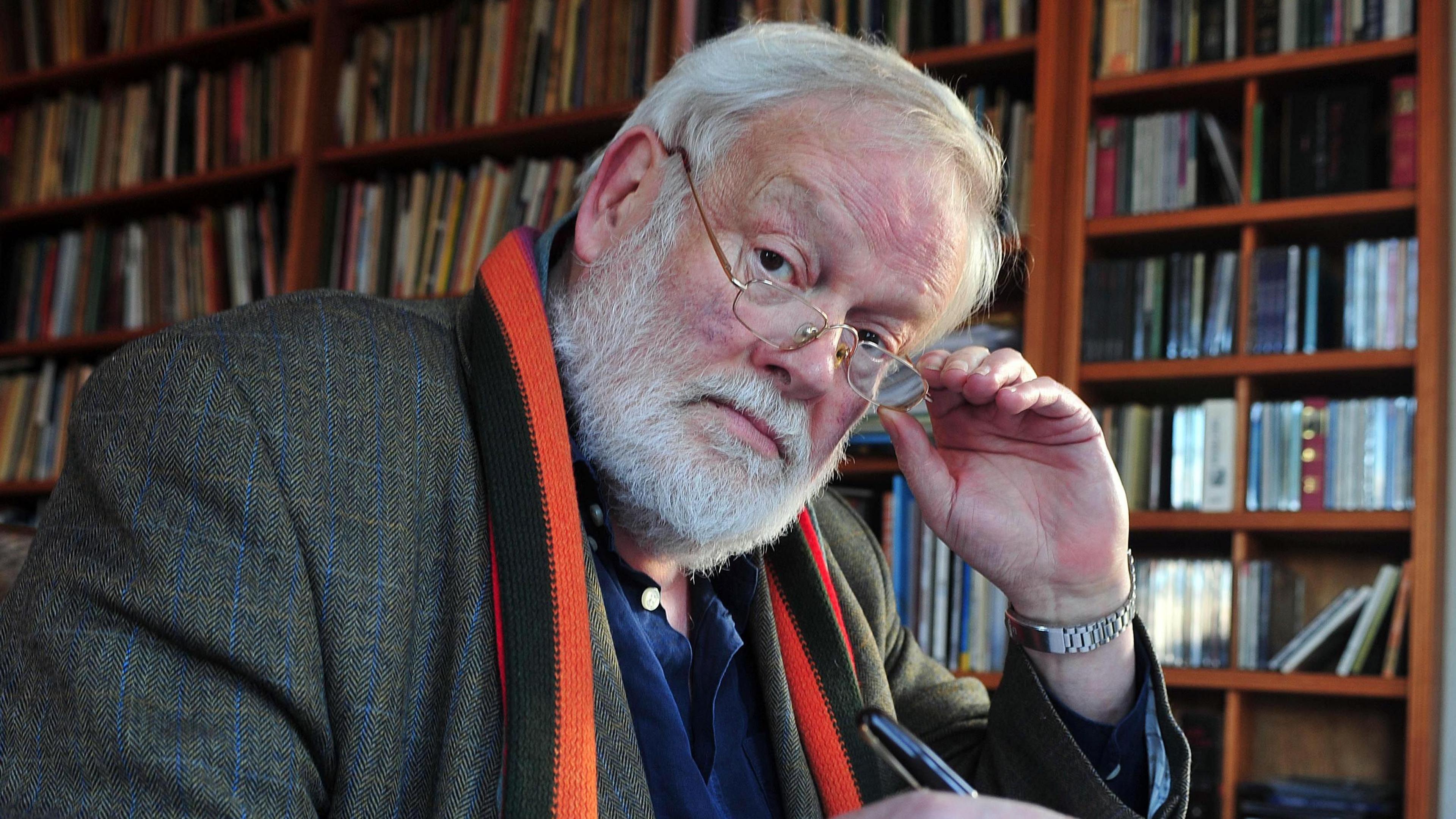 Michael Longley taking his glasses off in front of a brown book shelf, packed with books. He has white hair, a beard and moon shaped silver glasses. He is wearing a silver watch and a orange and black scarf. He has a grey tweed blazer jacket on. He is looking directly at the camera. 