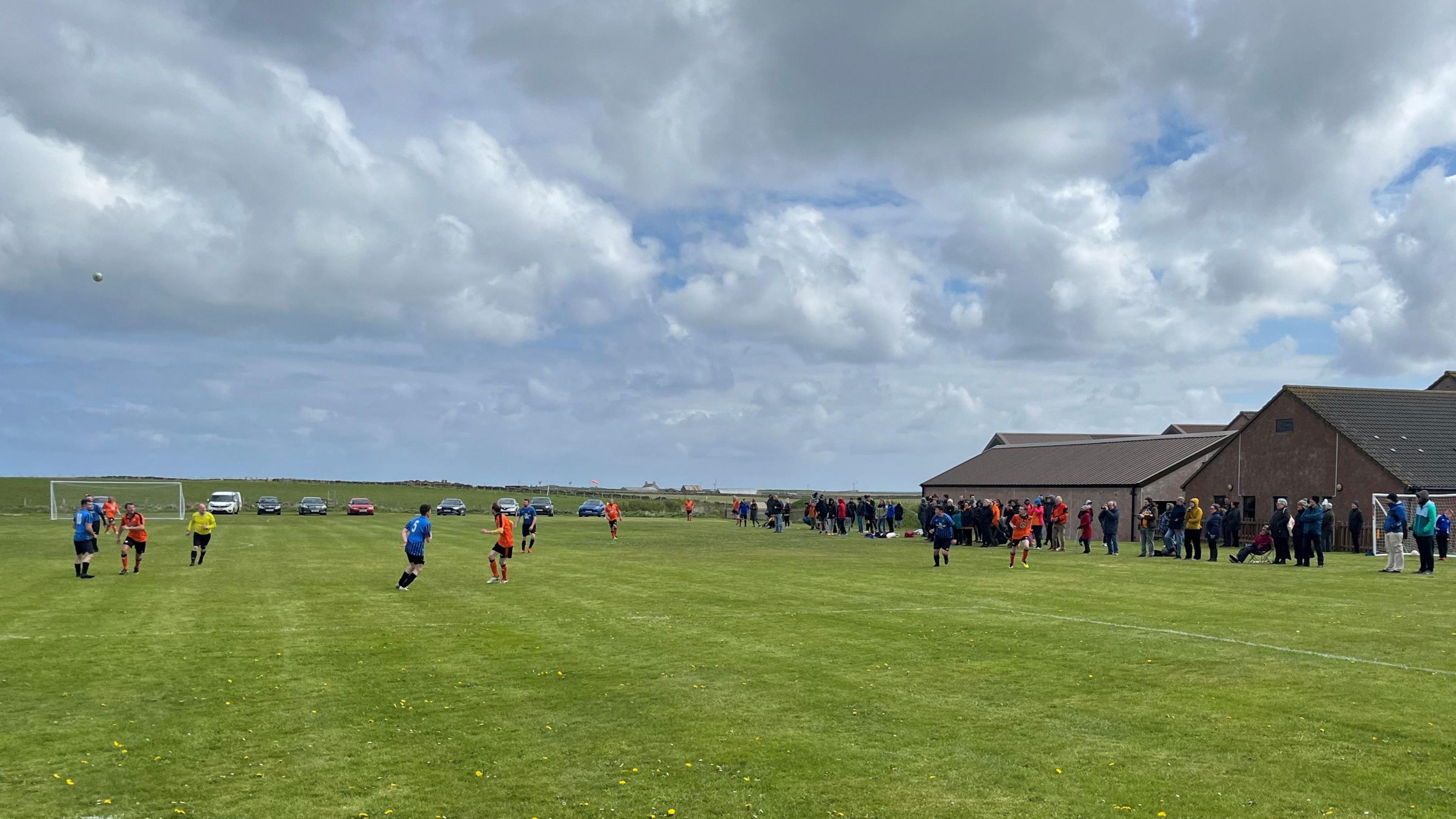 Men in orange and black playing football. Supporters watching at the side of the pitch.