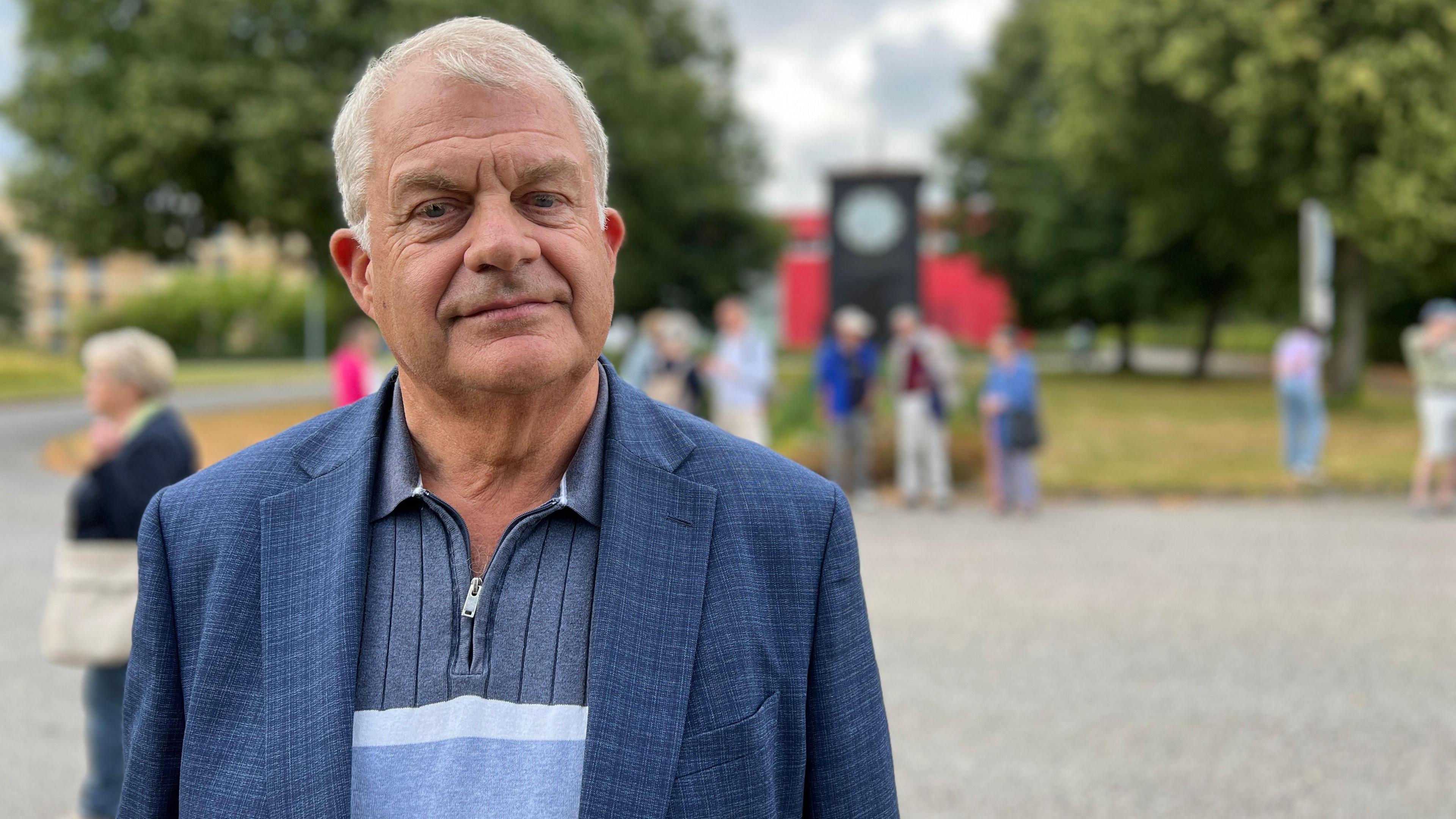 Sir Mark is standing outdoors, wearing a blue blazer over a striped shirt. In the background, several people are gathered in a park-like setting with trees and a large clock structure visible. The sky is partly cloudy