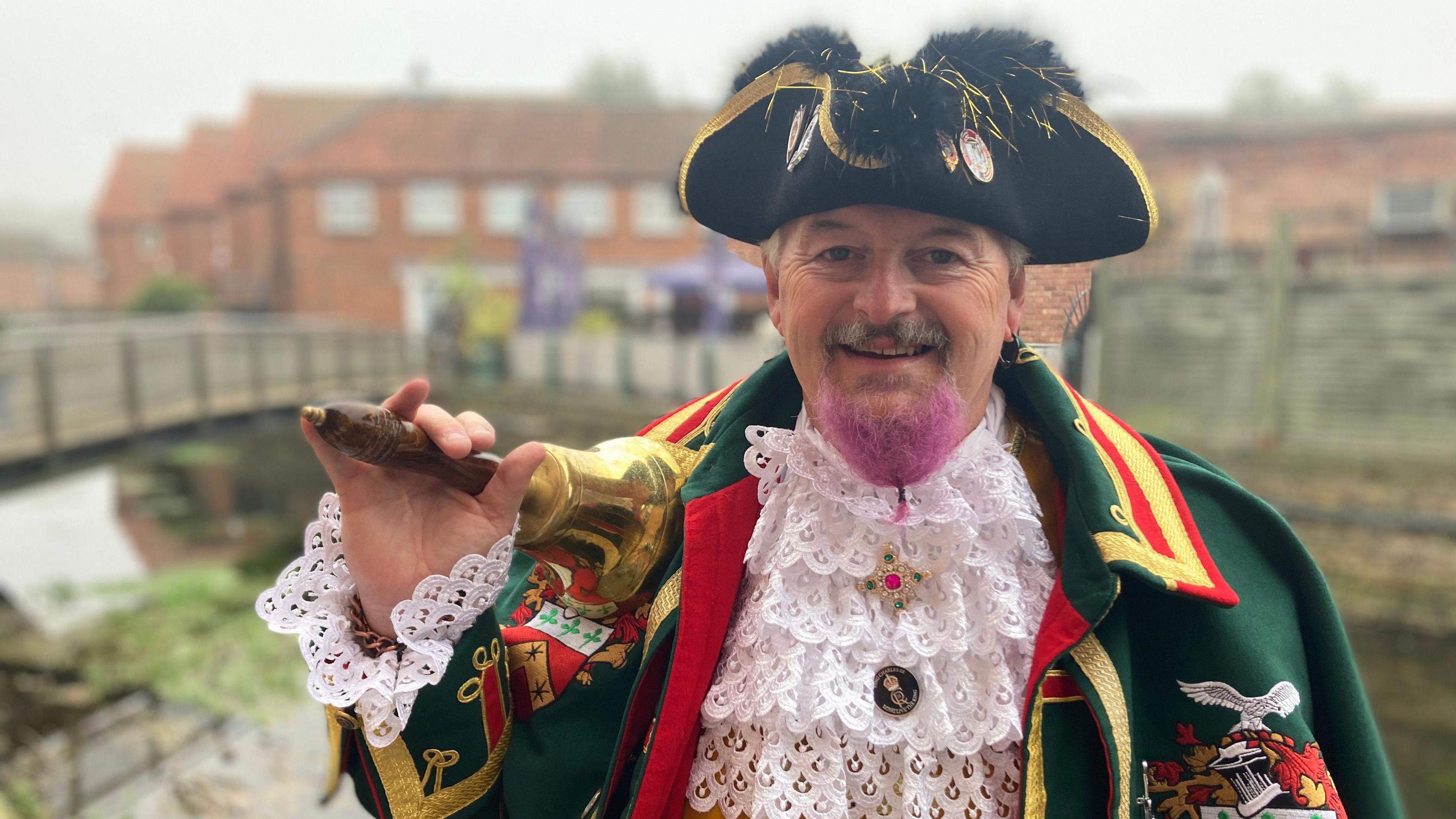 Sleaford town crier John Griffiths, dressed in a green regalia coat, a black hat with a gold rim. He is carrying a handbell and his beard is dyed pink.