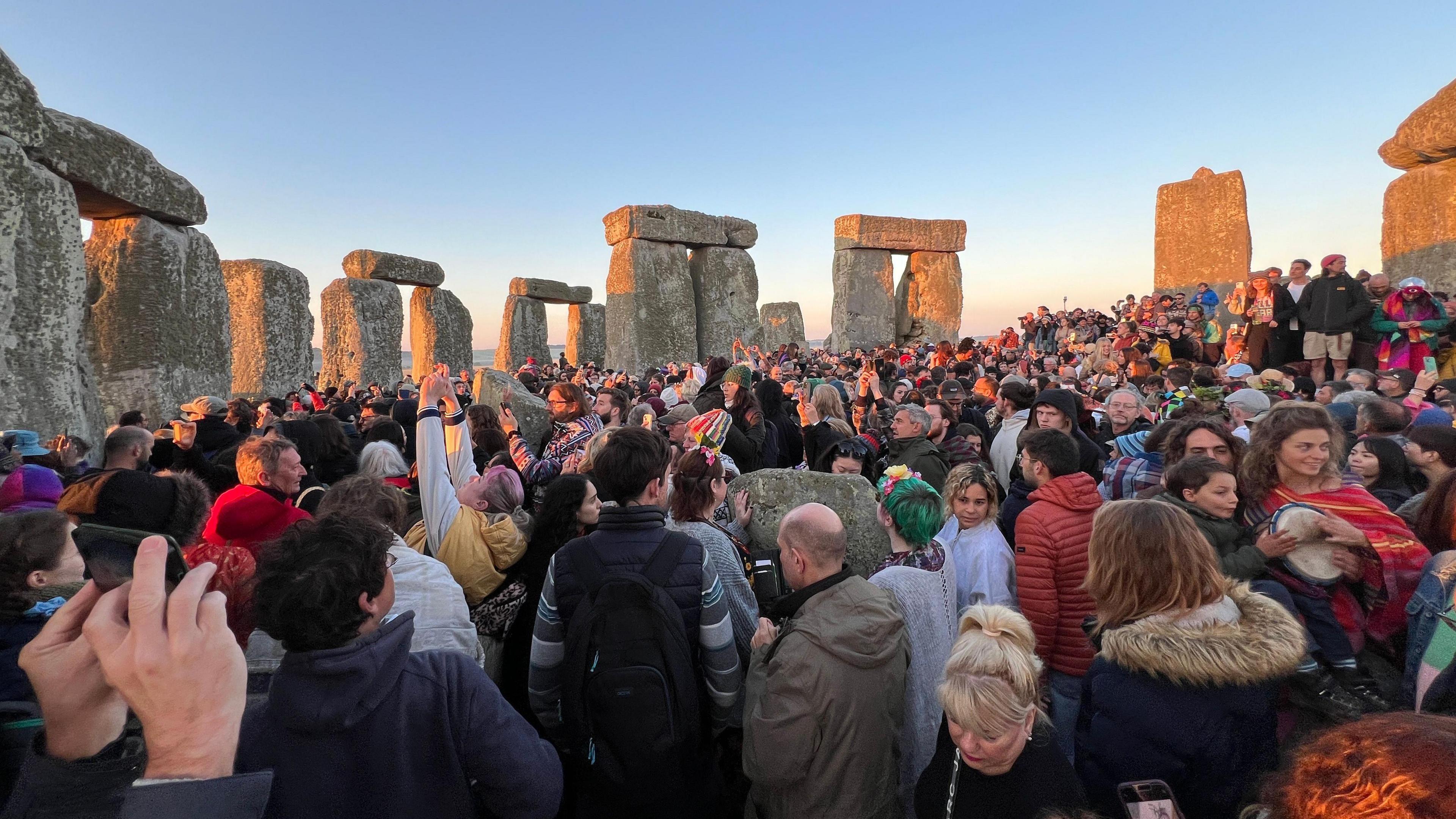 A large crowd of people gathered around Stonehenge as the sun rises over the horizon. The sky is light blue and one the left side of the stone sculptures are golden from the sunrise.