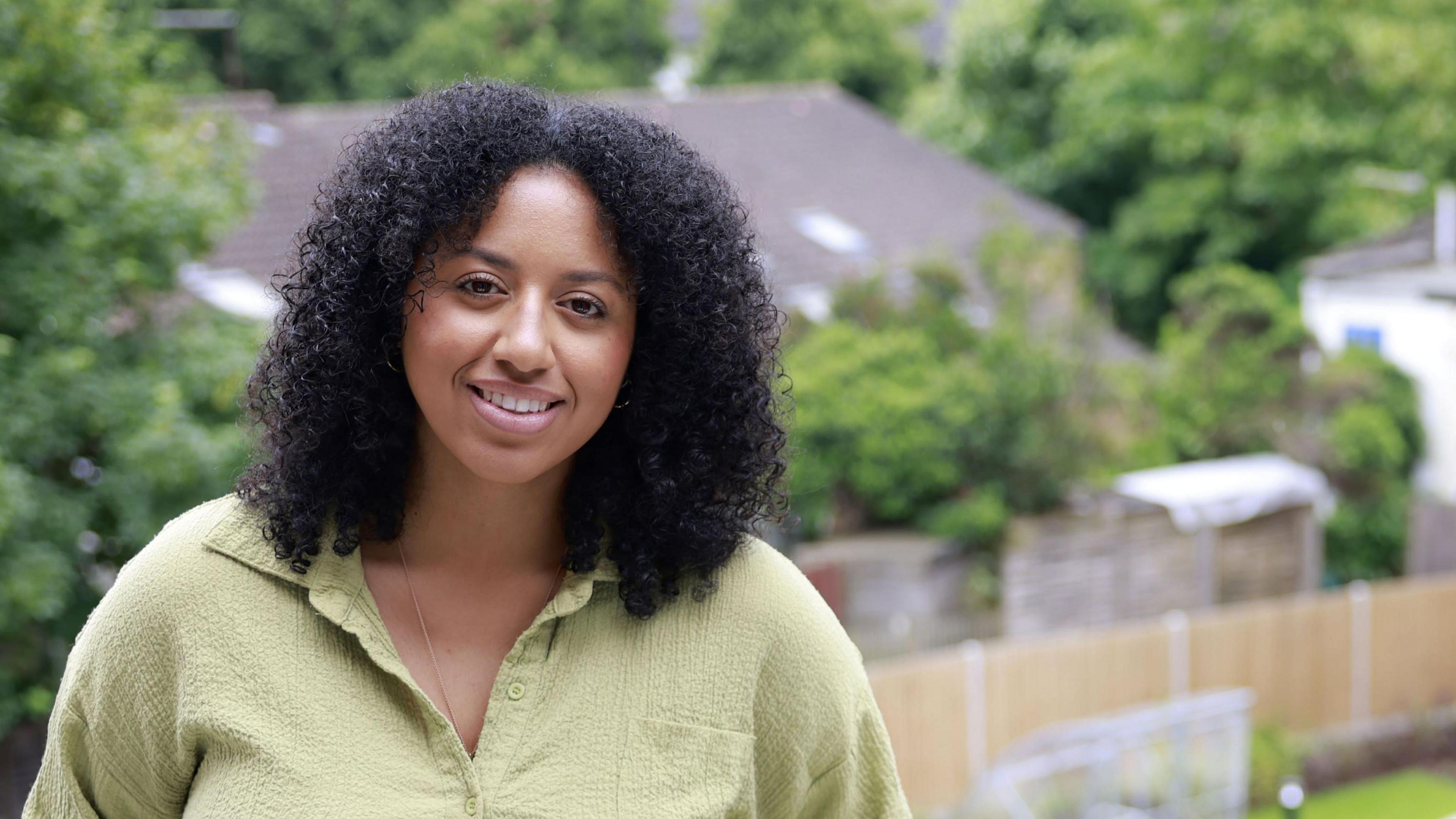 A woman with light brown skin and black, curly hair looking at the camera and smiling with teeth. She is wearing a green-yellow top. The background behind her is blurry, and shows a residential area with a back garden, trees and houses.