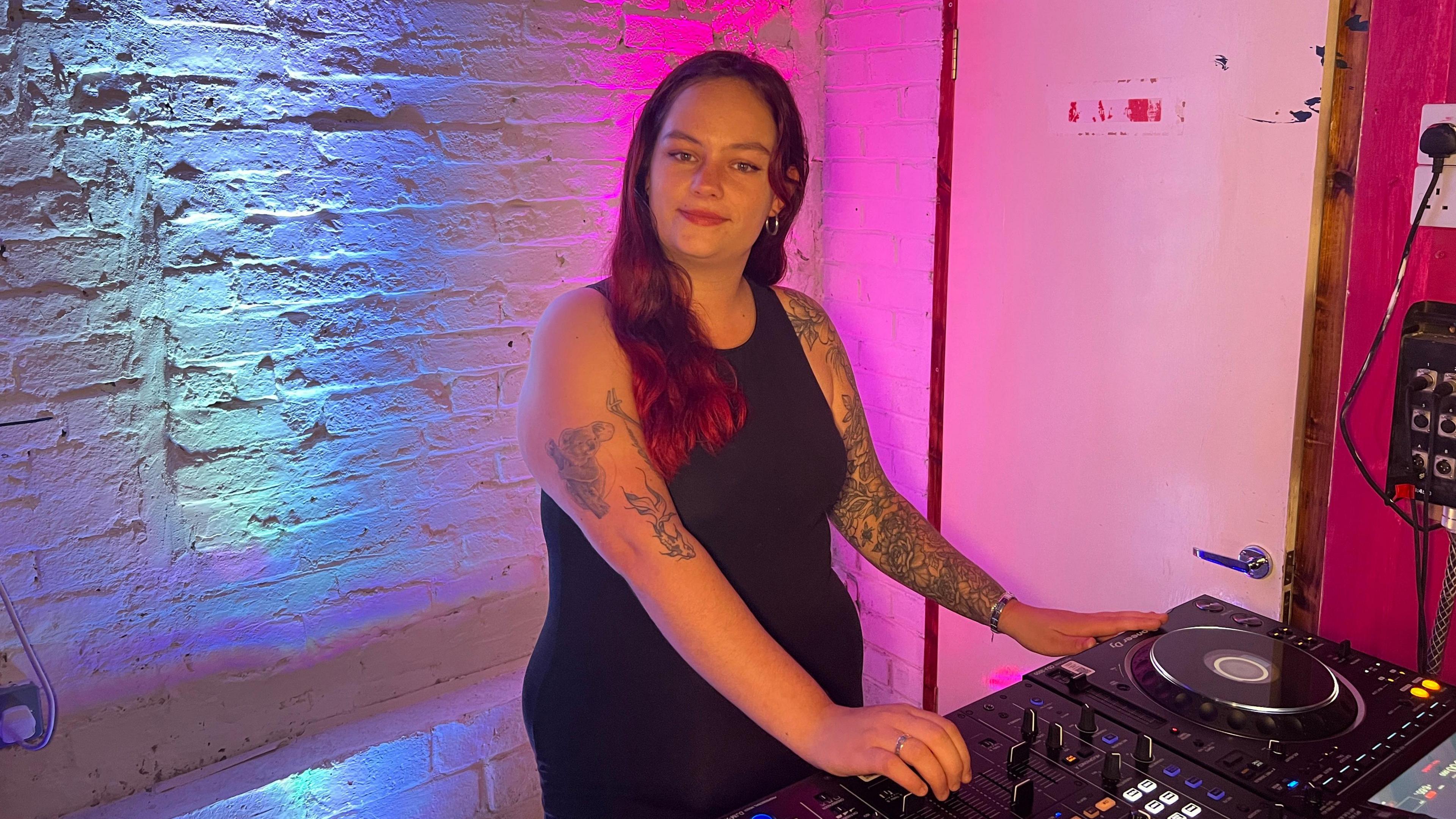 A white woman with long, red hair, stood over a set of DJ decks. She has a full sleeve tattoo on her right arm, and a tattoo of a fish and a koala on her upper left arm. She is smiling and is wearing a black vest and black trousers.