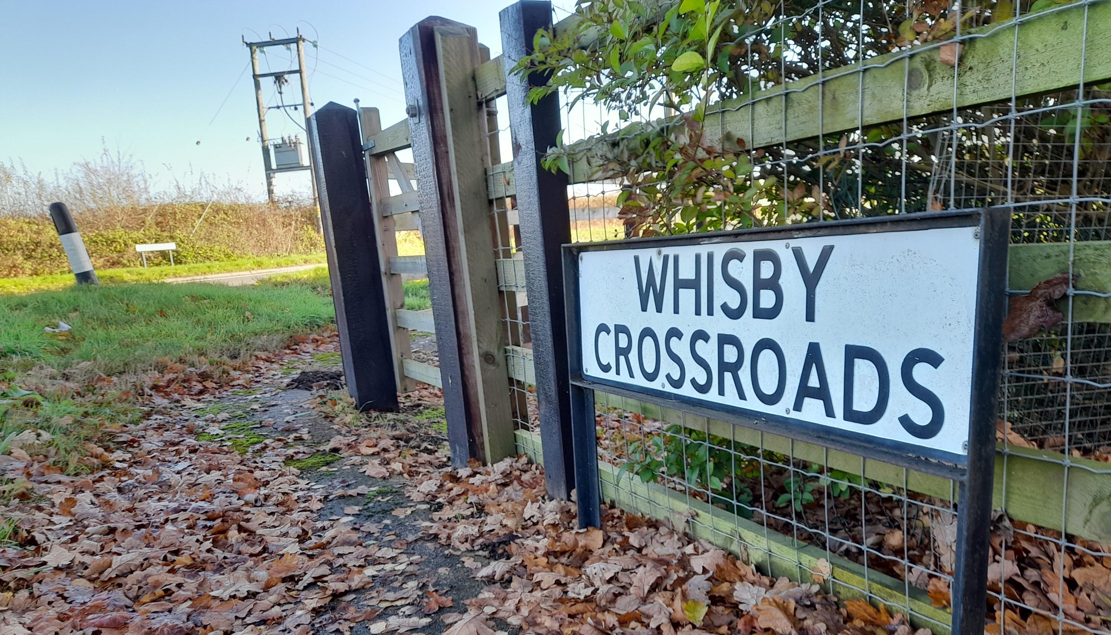 A sign on a fence which reads "Whisby Crossroads". The road is in the background and there are leaves along a path