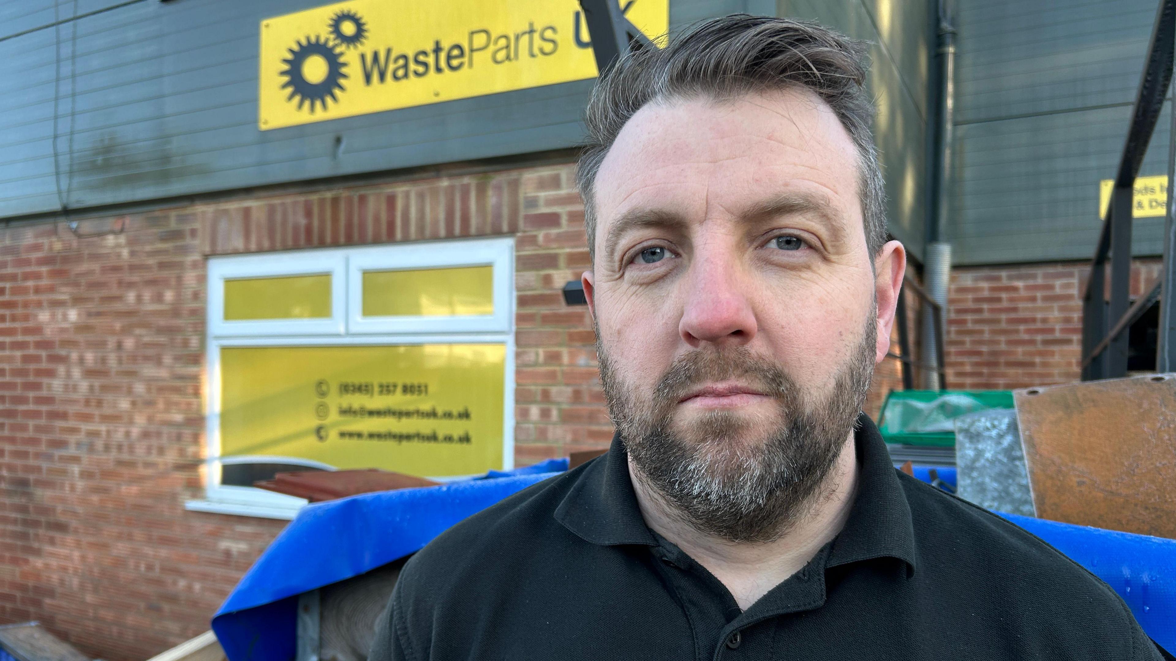 Tyrone Behan outside his warehouse. He has a beard and is wearing a black polo T-shirt. There is yellow sign behind him saying WasteParts UK. 