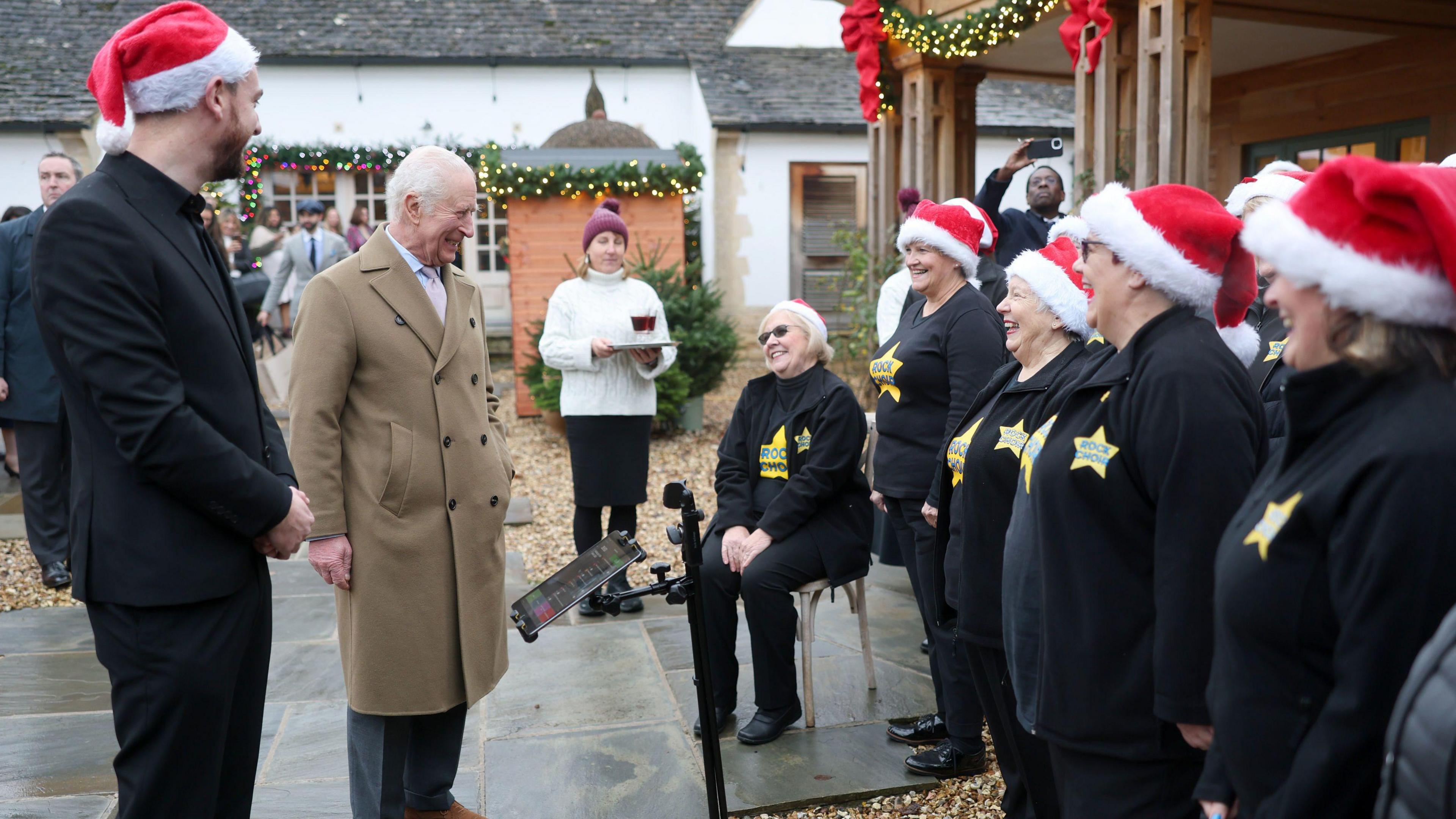 King Charles III meeting carol singers. He is wearing a light brown coat over a white shirt and pink tie. The carol singers are all in black jumpers with a star on the side. They are also wearing red santa hats. The King is looking at the singers and they are all smiling. 