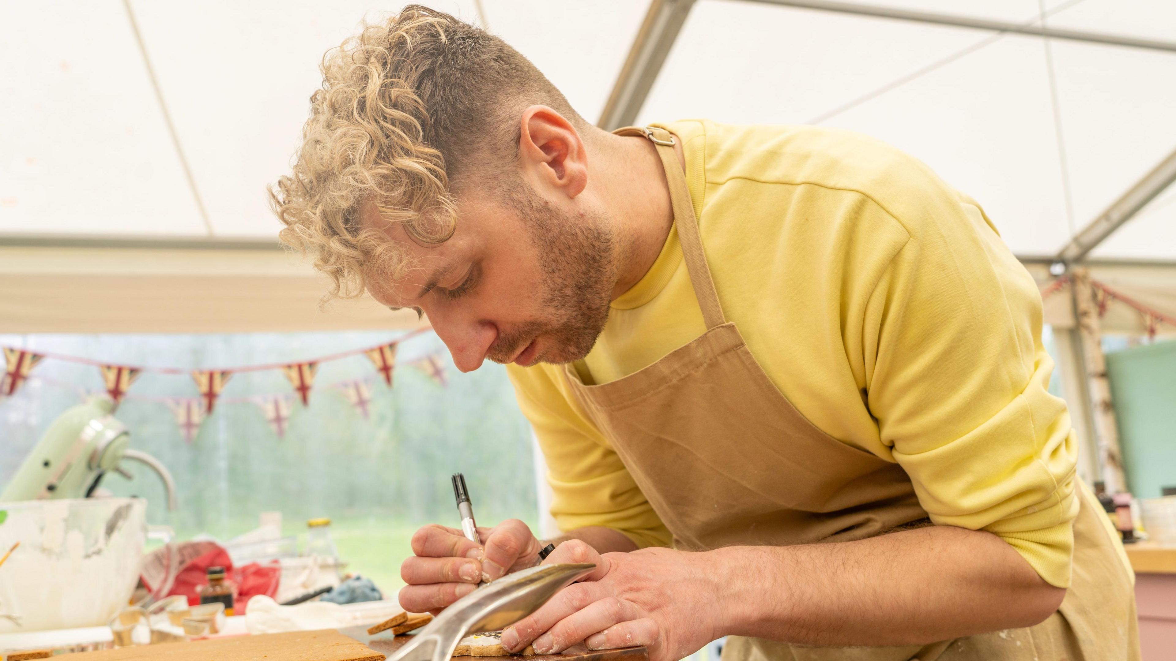 John Mincher has blonde hair and is wearing a yellow top. He is working on a cake creation in the Bake Off hut. There is bunting in the background with a Union Jack print.