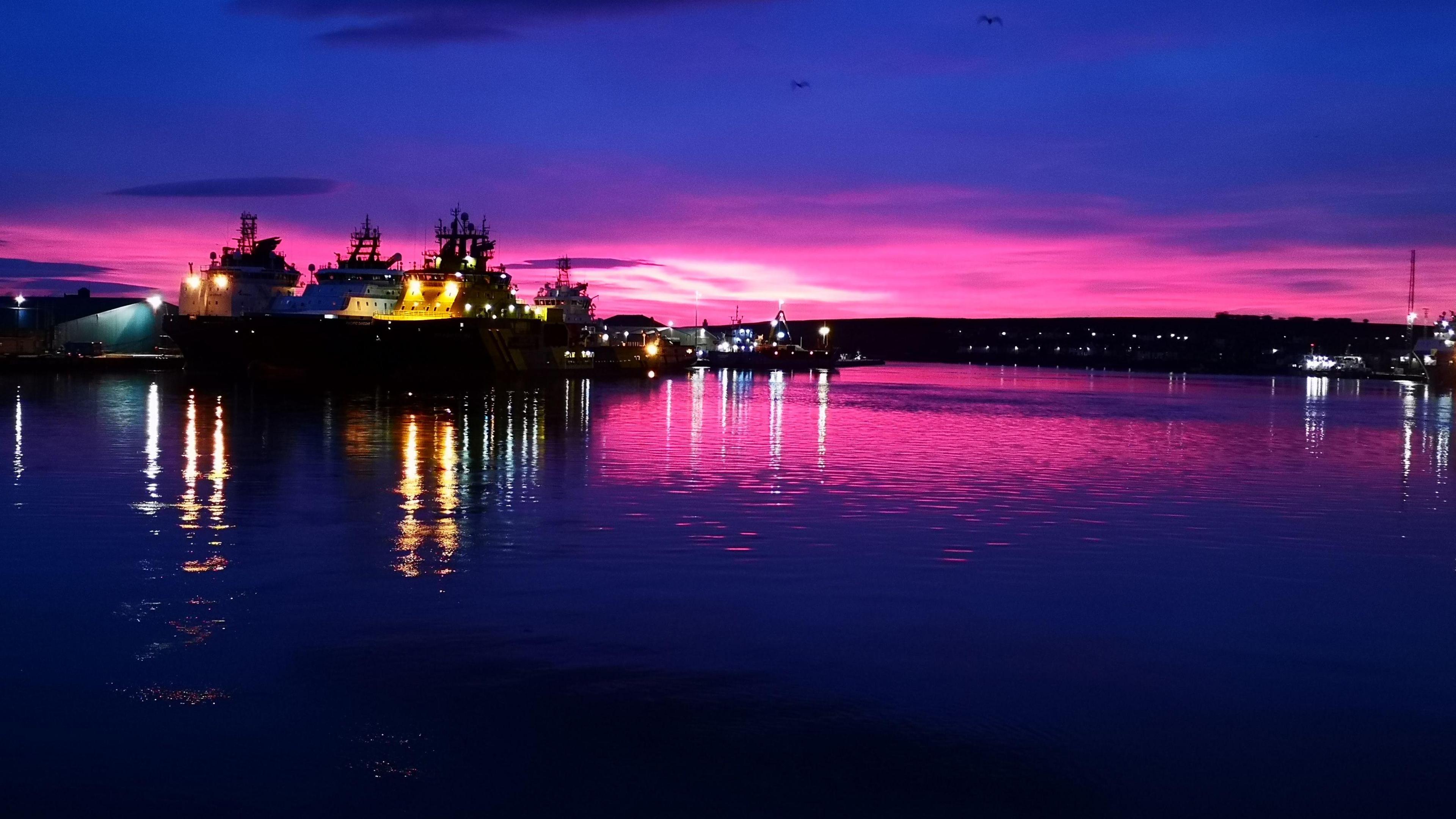 A harbour at night with a dark blue and pink sky and the lights from a number of boats shining out and reflected in the water