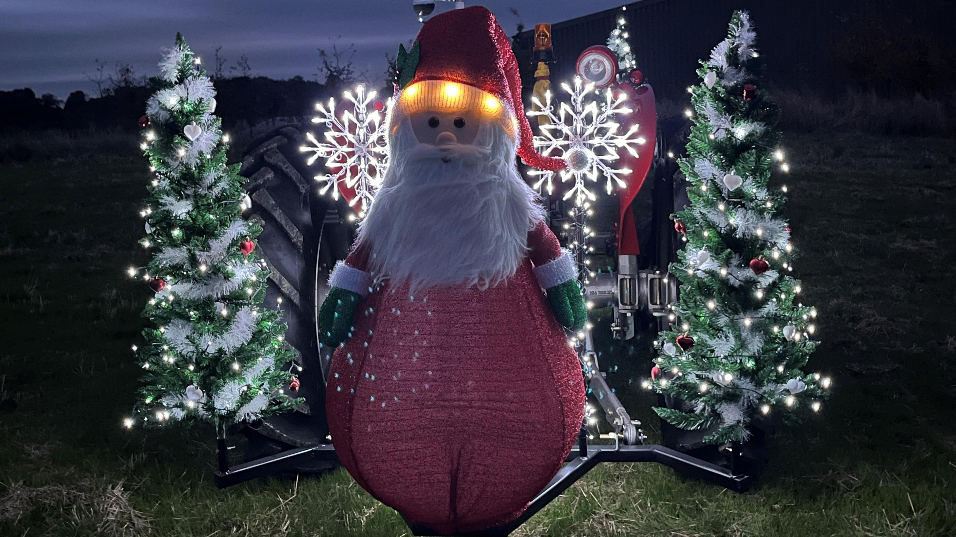 A model of a Father Christmas sits at the front of a tractor and Christmas trees are attached to either side. It is lit with fairy lights.