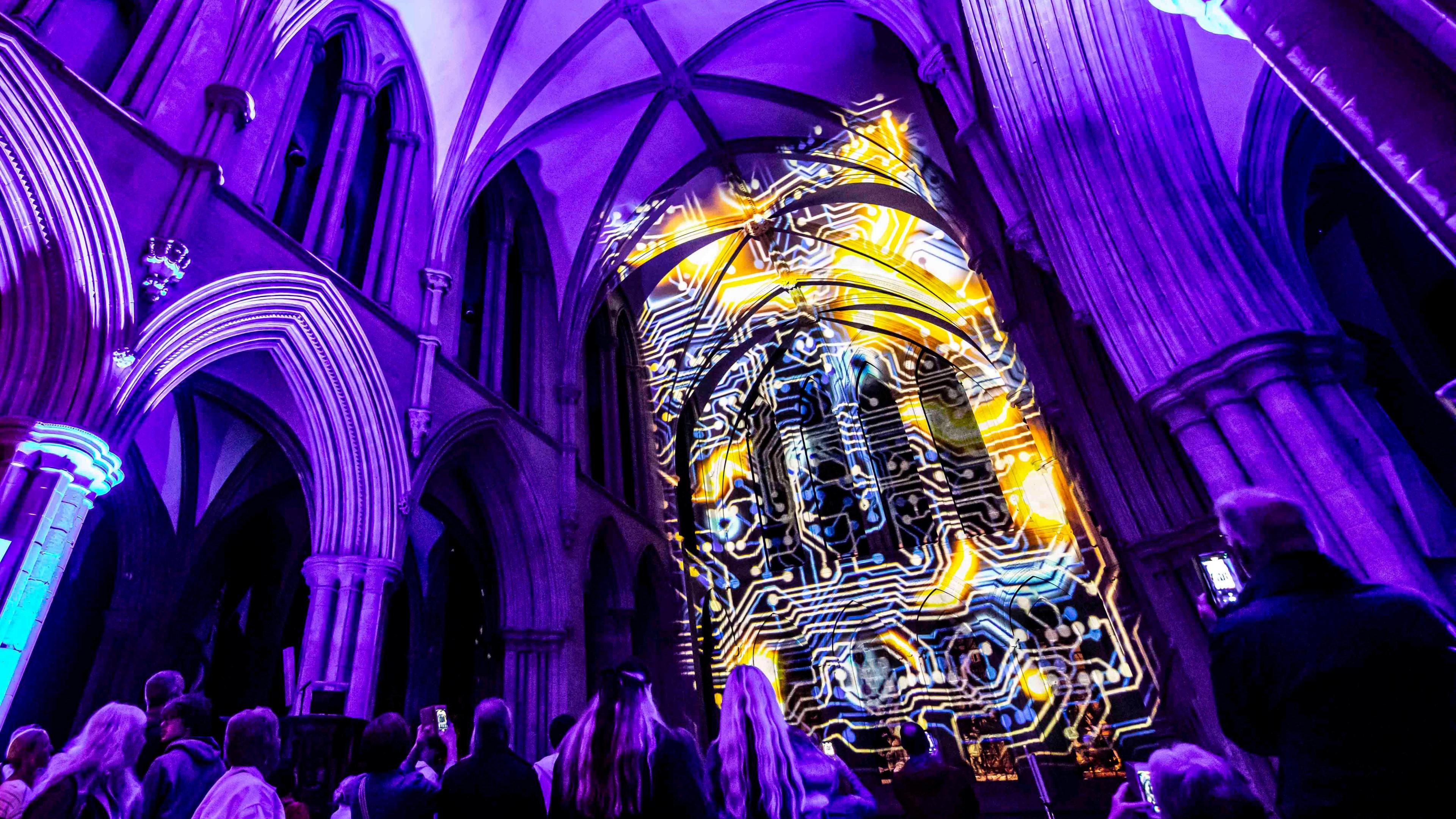 A light projection against the walls of a church. People are seen from the back looking at the installation.