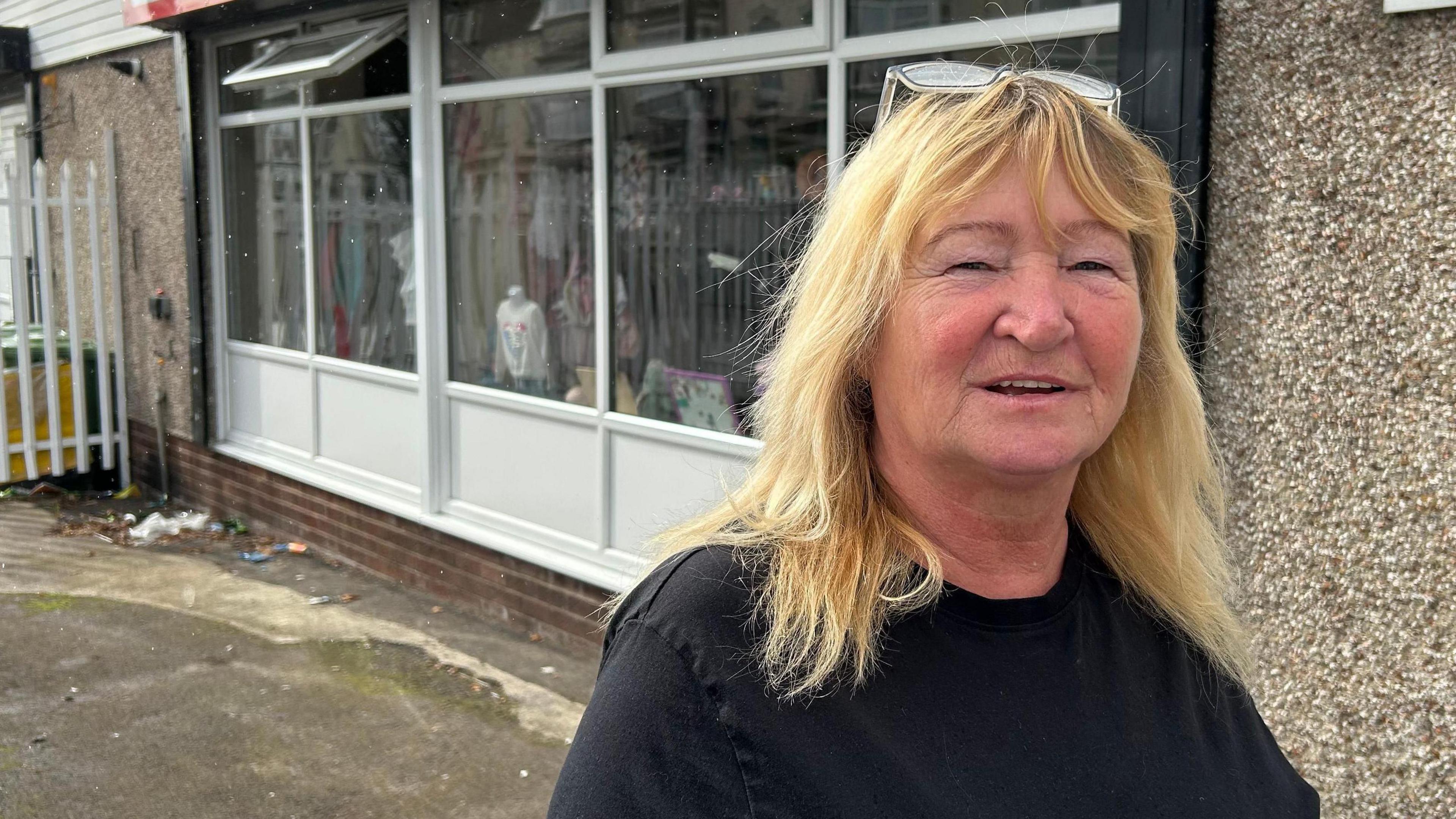 Wendy English, the founder of Second Chance Sunderland, stands in front of its new store in Hendon. The shop has big glass windows with white wooden frame and the wall is covered with pebbledash. She has shoulder-length blonde hair and a fringe, with her glasses on her head, and is smiling. Her jumper is navy blue.