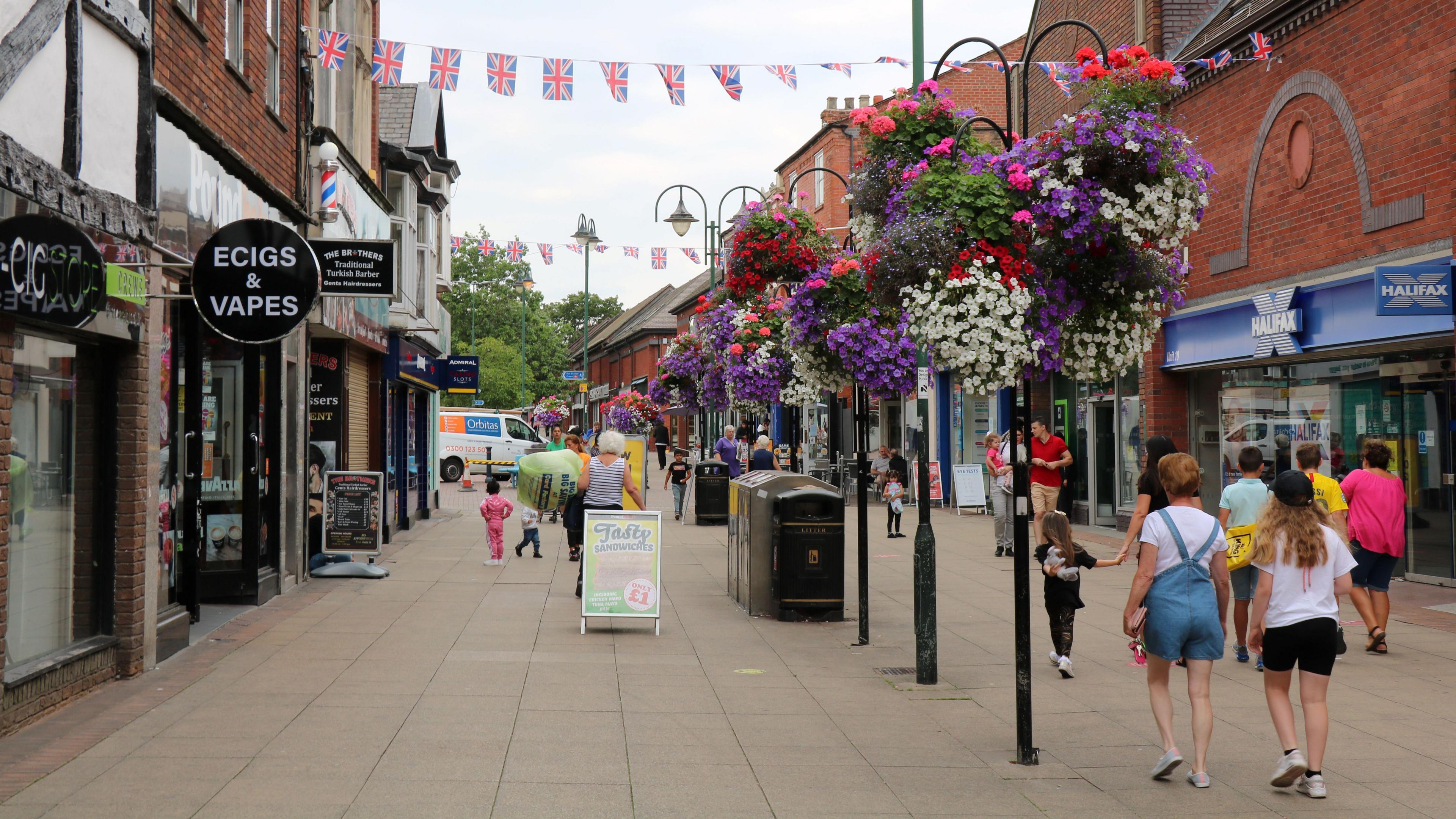 A town centre shopping street with people walking and hanging baskets decorating the centre of a paved area, there are Union Jack flags flying and shop fronts either side of the image.