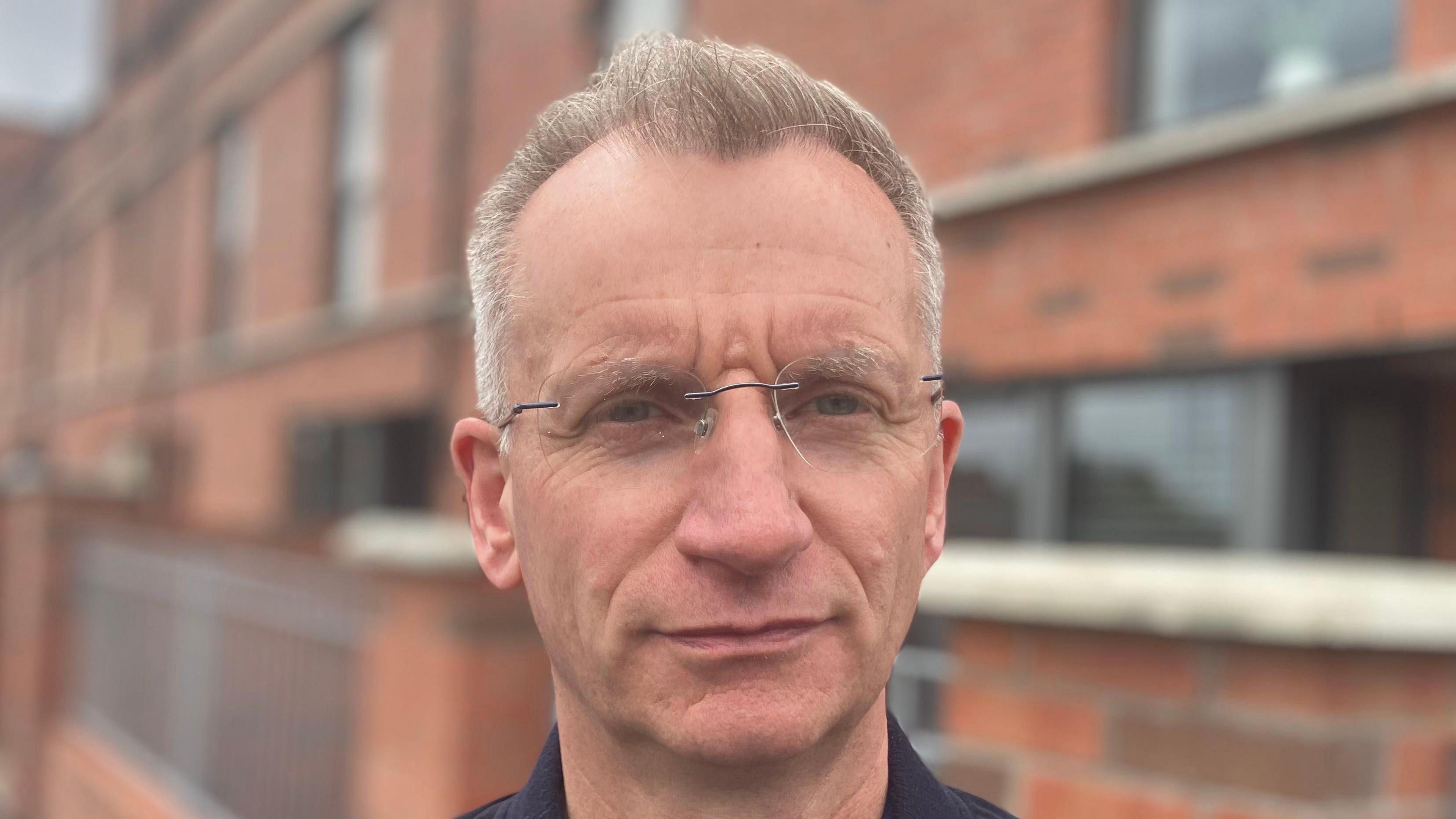 Kingston has short grey hair and eyebrows, he is wearing clear and metal rounded glasses. He is pictured in a close-up shot and is wearing a black jacket.