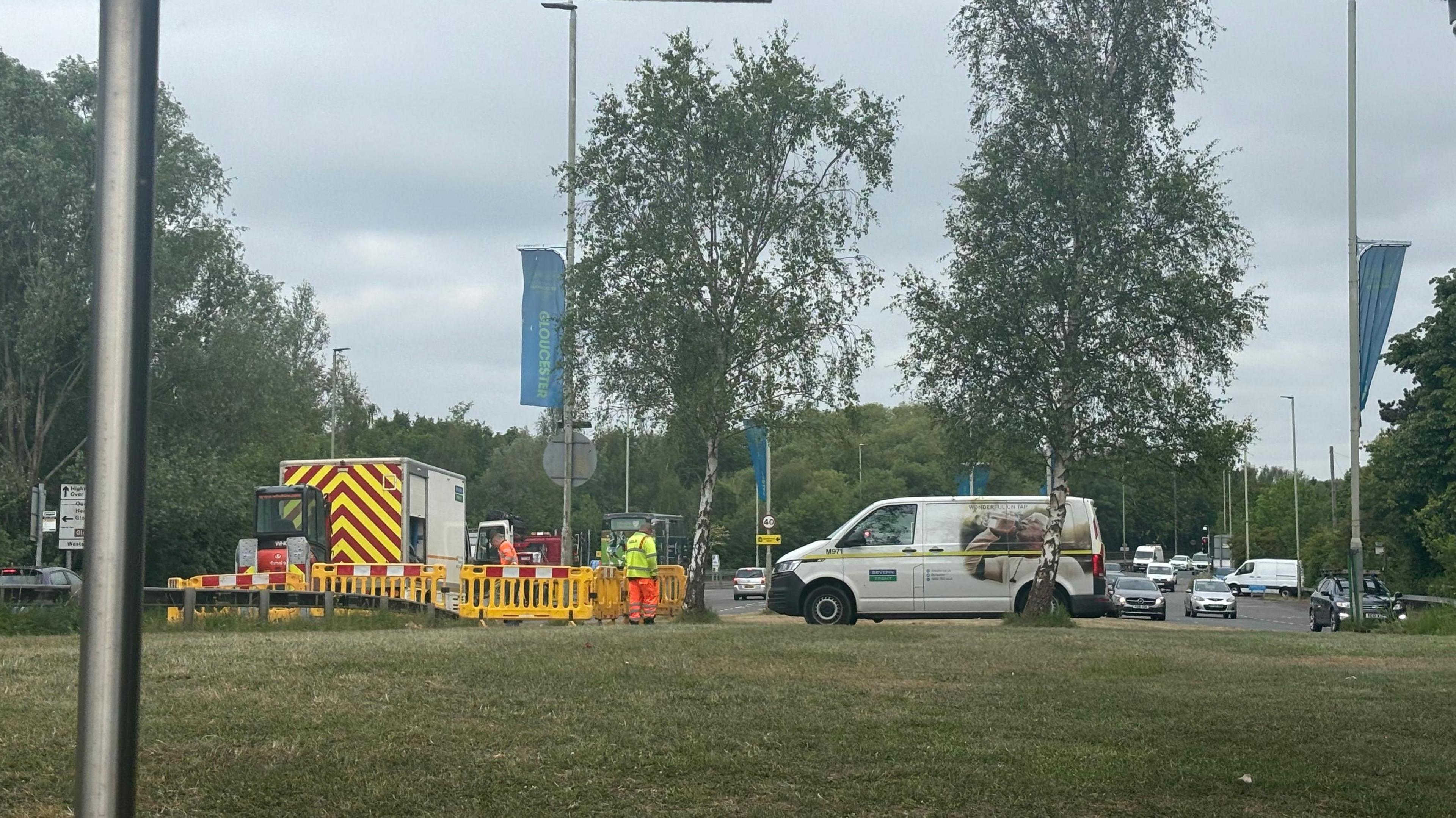 A team of two men from Severn Trent, wearing hi-vis clothes, stand near parked vans on a roadside grass verge near a busy road.