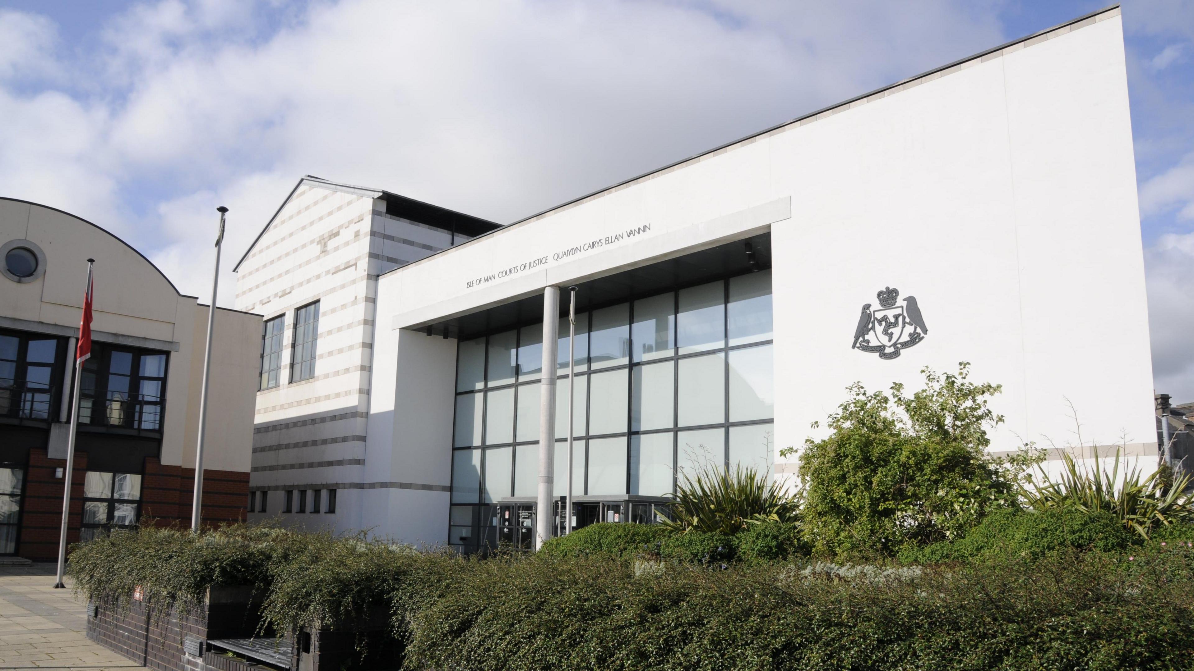 A modern courthouse building, with white walls and the Isle of Man Government Crest, and a large glass entrance. There are hedges in planters next to benches that are built into the walls in the foreground. 