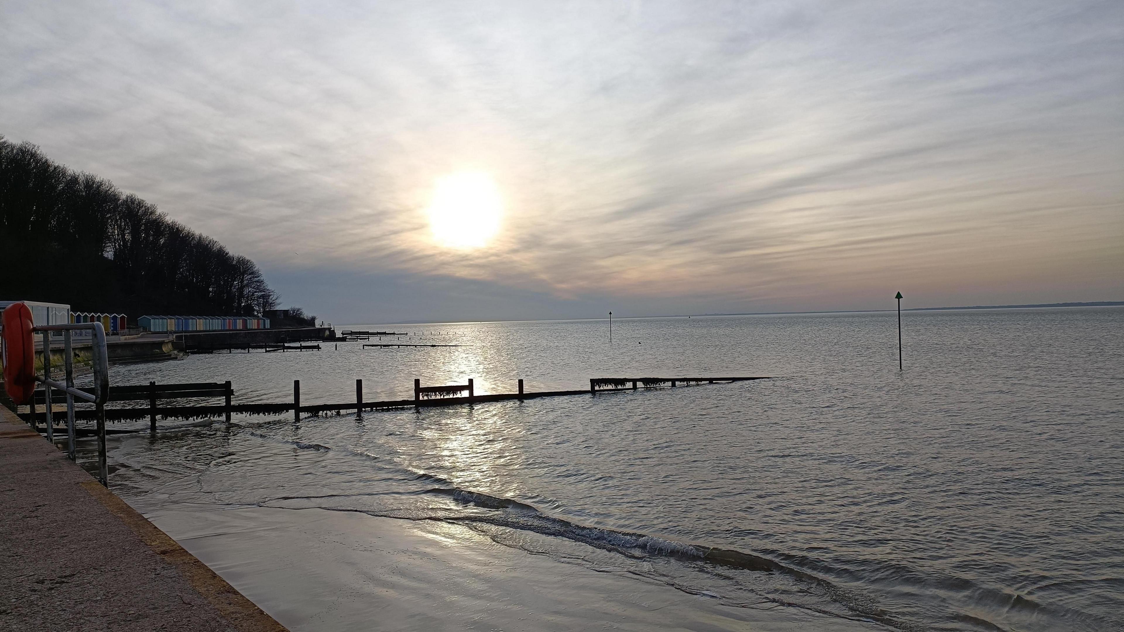 Sunset over a beach scene. The sun is a yellow haze in the sky with a few wispy clouds. The beach is deserted with a few beach huts seen in the distance.