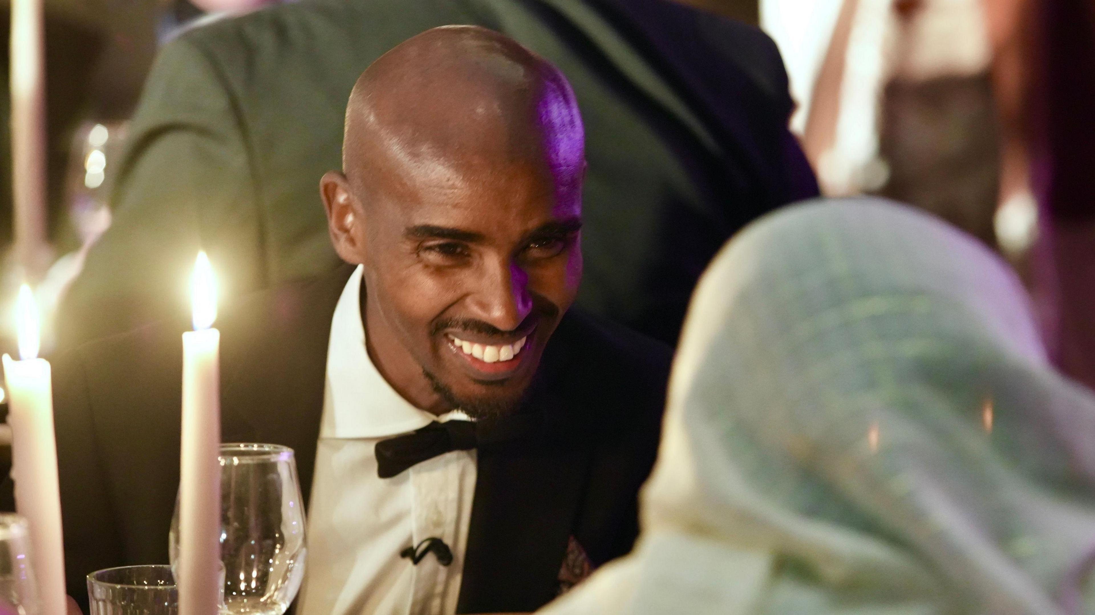 Sir Mo Farah smiles as he chats to another guest during an event at The Mount Without in Bristol. He is wearing a black bow tie and dinner jacket and his face is partially illuminated by two candles