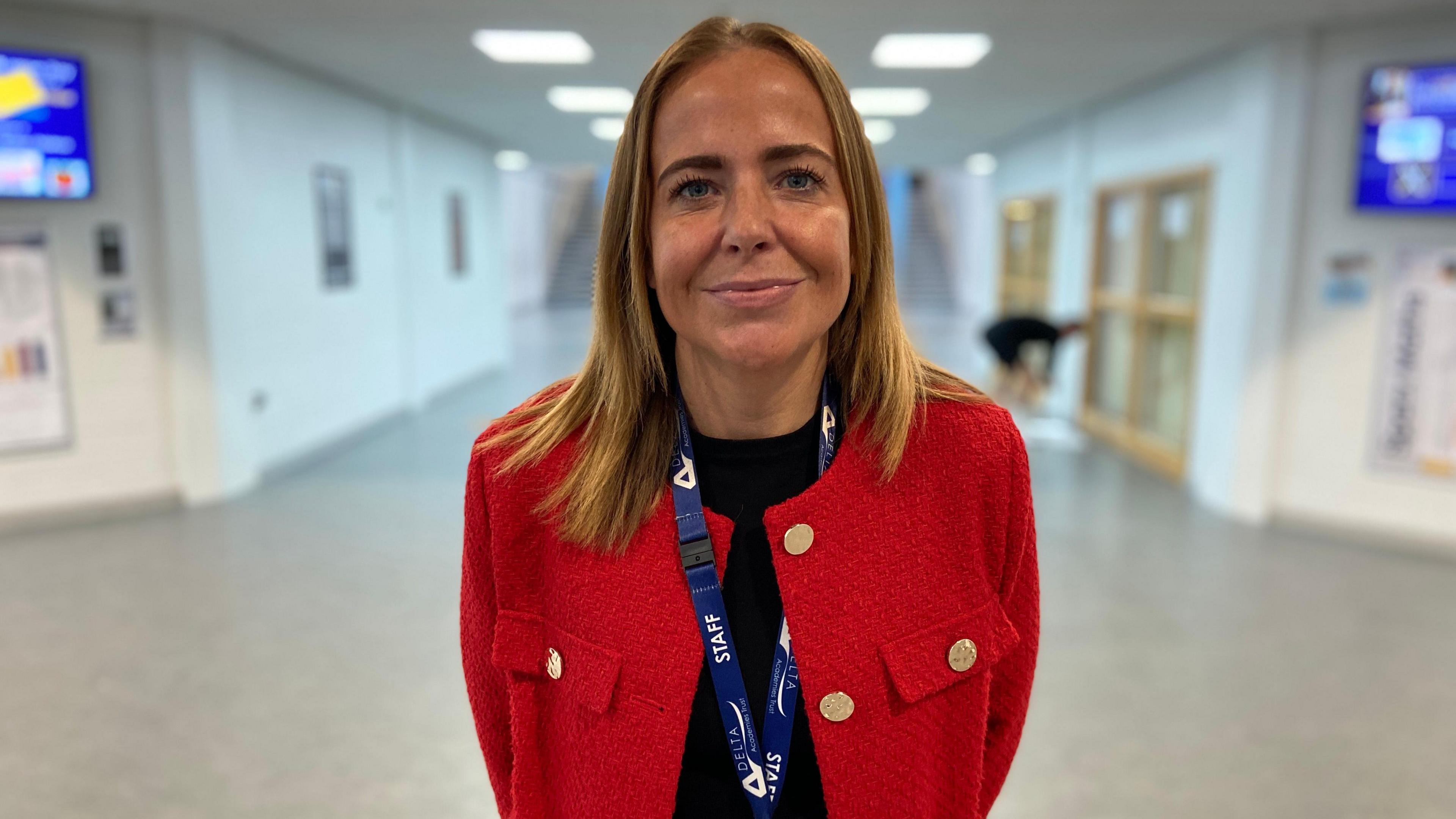 A head teacher stands in a bright, white atrium of the school hall. She has shoulder-length blonde hair, wears a bright red suit jacket and is smiling at the camera.