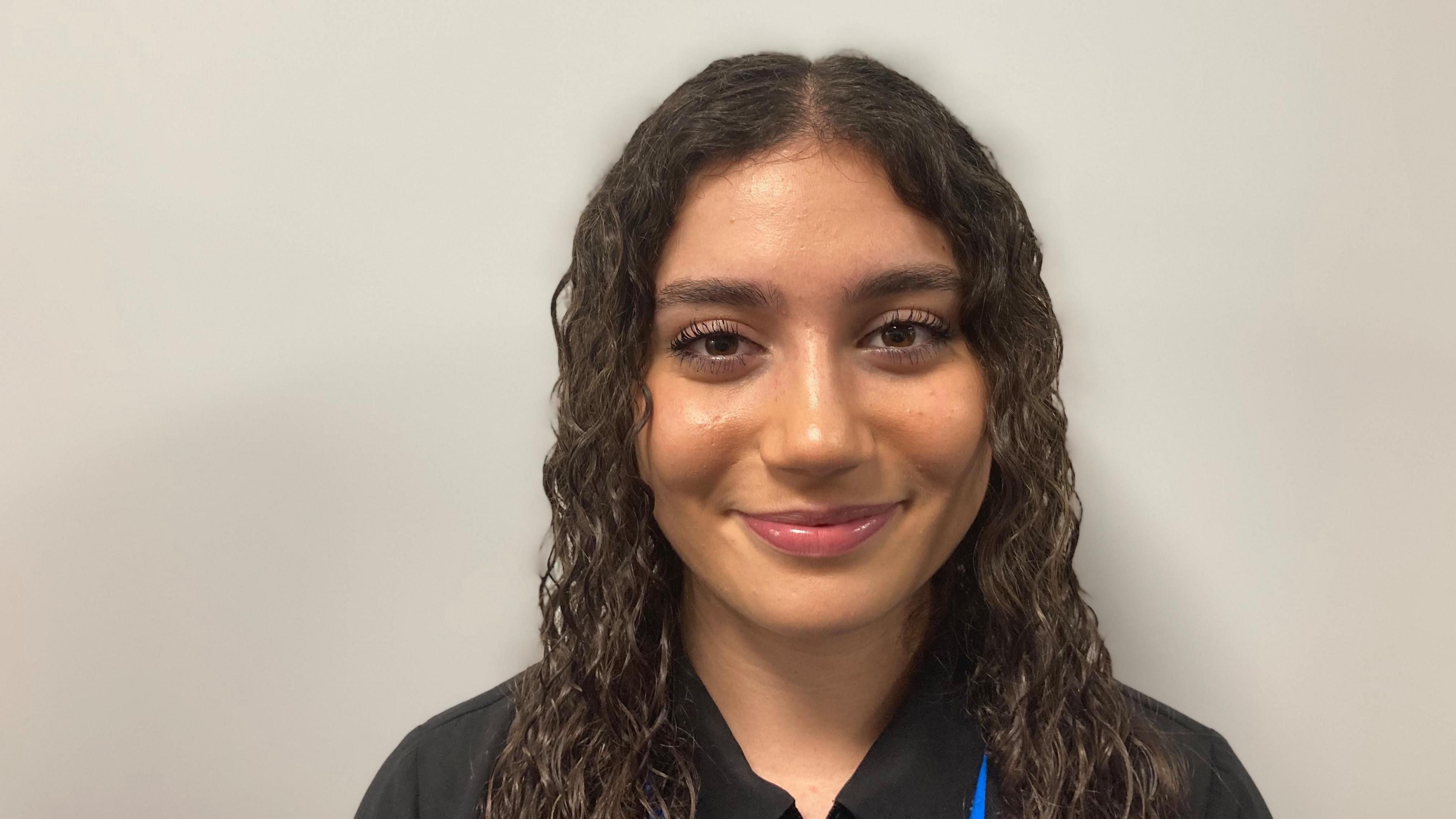 Deniz smiles at the camera, she wears a black shirt and has long black hair.